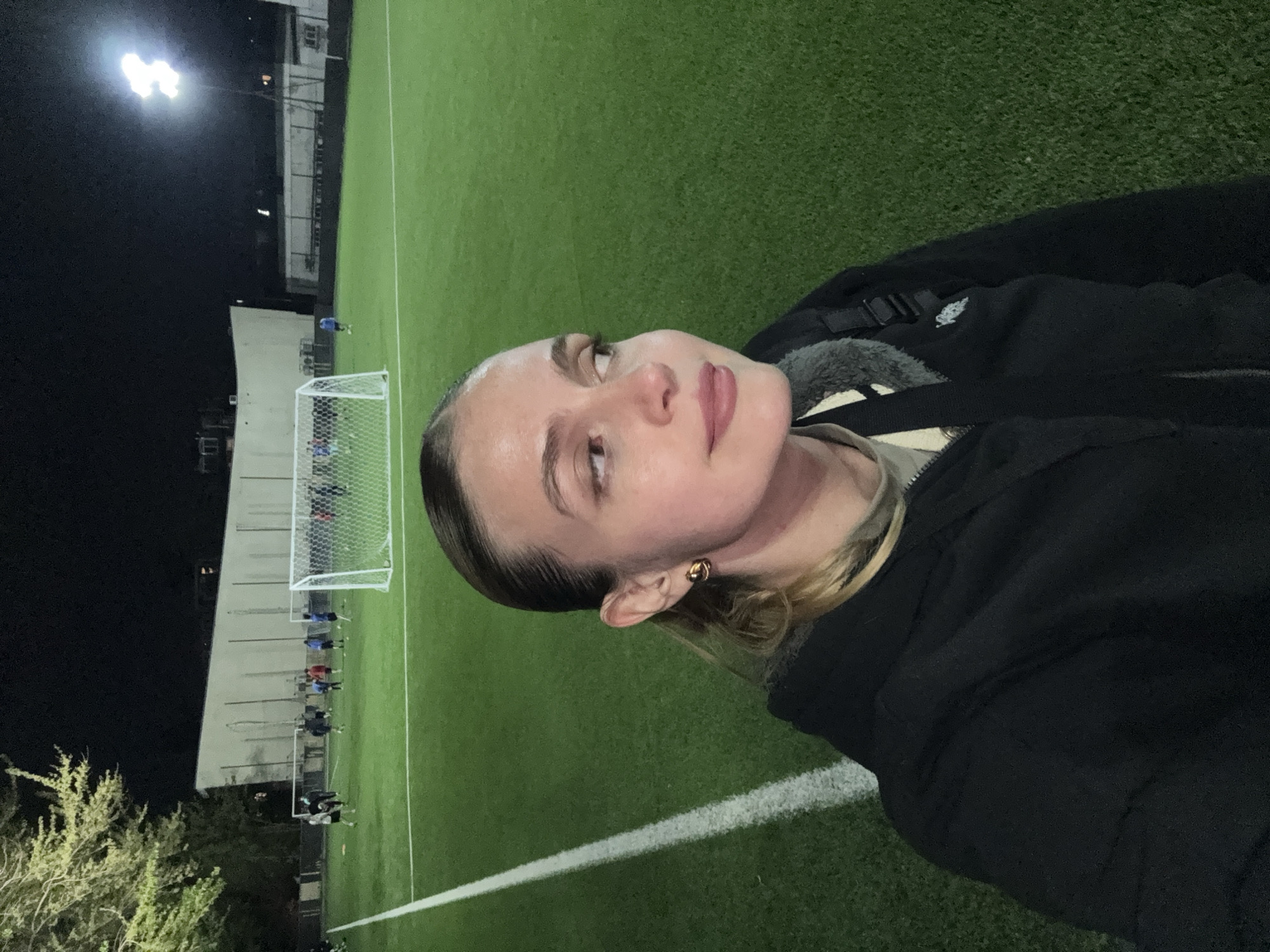 The image, taken at night in Tbilisi, Georgia, features a young woman in the foreground taking a selfie or being photographed on the sidelines of an artificial turf soccer field. She has light skin, dark eyebrows, and light-colored hair, some of which is pulled back, while blonde strands are visible near her right ear. She wears a gold-colored hoop earring in her right ear and is dressed in a dark, possibly black, jacket with a grey or white fluffy lining visible around the collar. Her expression is calm and neutral, as she looks slightly upwards and to her left.
The setting is an outdoor sports complex, illuminated by bright floodlights visible in the upper left. The green artificial turf field, marked with white lines, extends into the background. On the far side of the field, a white soccer goal is visible. Several figures, appearing to be players or spectators, are gathered near the goal, suggesting a game or practice is underway. In the deeper background, dark silhouettes of buildings and some trees are discernible against the night sky. The scene is lit by artificial light, indicating it is nighttime. No discernible text is visible in the image.