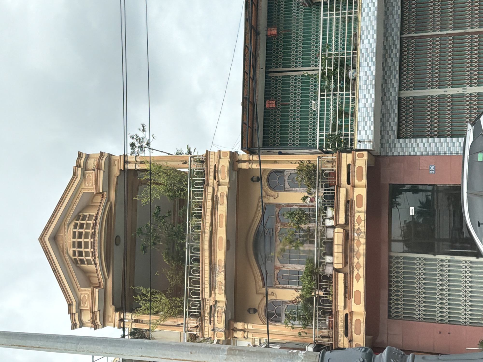 A medium shot captures two adjacent buildings in Phú Xuân Ward, Vietnam. The building on the left is a narrow, multi-story structure painted in a light yellow and pink. The upper floors feature balconies with ornate railings and potted plants, while the lower floors have large, segmented metal gates and a glass door. The building on the right is shorter and wider, with a green metal gate covering its facade. A number '396' is visible on a square plaque on the pink section of the left building. Overhead, power lines stretch across a cloudy sky. The scene is devoid of people and depicts a street view of urban architecture.