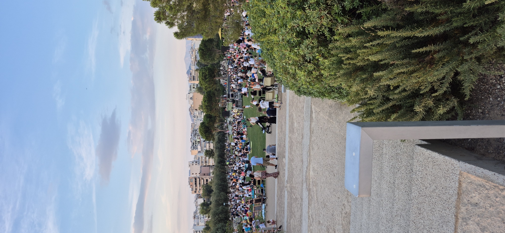 This image captures a lively outdoor event at Stavros Niarchos Park in Kallithea, Greece. A large crowd of people is spread across a grassy area, many seated on blankets and chairs, enjoying what appears to be a performance on a stage in the middle ground. The stage has a black backdrop and is surrounded by lights and equipment. Buildings and trees form the background, with a hint of mountains in the far distance. The sky above is a soft blue with scattered clouds, indicating it is either early morning or late afternoon. The foreground shows a paved area with concrete structures and a dense green bush on the right. The atmosphere suggests a relaxed and communal gathering, likely a concert or cultural event.