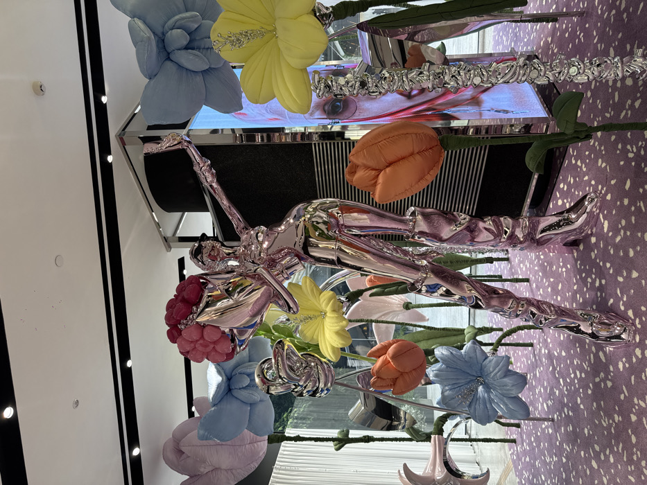 The scene depicts a vibrant, artistic installation within a luxury retail environment in Bangkok, Thailand. The central subject is a large, highly reflective, chrome-pink humanoid sculpture, appearing female, with stylized, angular features. The figure strikes a dynamic pose, holding what appears to be a smartphone in its extended right hand, simulating taking a selfie. Its left arm cradles a bouquet of bright pink, globular flowers. The sculpture exhibits wing-like structures on its back and wears ornate, high-heeled boots that crumple around the calves.
Surrounding the central figure are numerous oversized, soft-sculpture flowers made of fabric. These include large light blue, pale pink, and yellow lotus-like blooms, as well as orange tulip-shaped flowers, all supported by thick, green, fabric-wrapped stems. Interspersed among these are slender, twisted, metallic silver structures resembling abstract plant forms. The floor is covered in a speckled purple carpet. In the background, large mirrored pillars and digital display screens reflect parts of the installation and a blurred outdoor or internal garden scene visible through a large window or glass facade, suggesting an open and modern architectural space. Overhead, white ceilings feature recessed lighting. No actual human figures are visible.