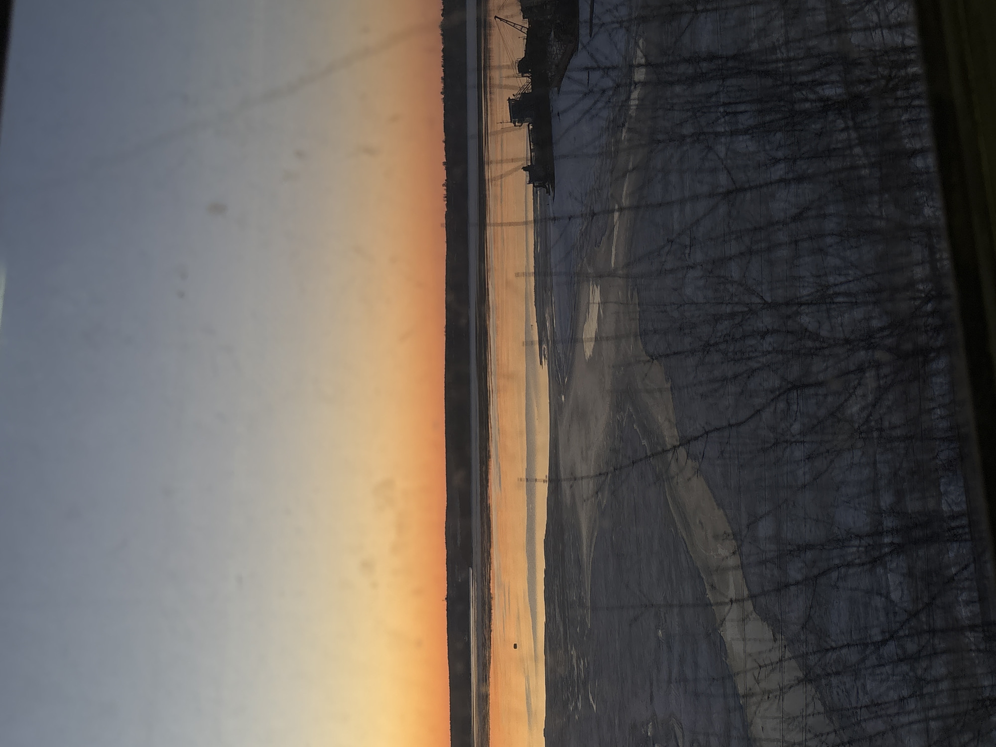 The image shows a sunset view over the Kama River in Perm, Russia. In the foreground is an open window, through which the landscape is viewed. The left side of the image depicts the sky transitioning from blue to orange as the sun sets. The Kama River, a significant waterway, dominates the center. On the right is a dense area of trees. In the distance, outlines of structures or industrial installations can be seen along the riverbank. The image is captured through the window, with what appears to be a reflection of tree branches overlaying the view.