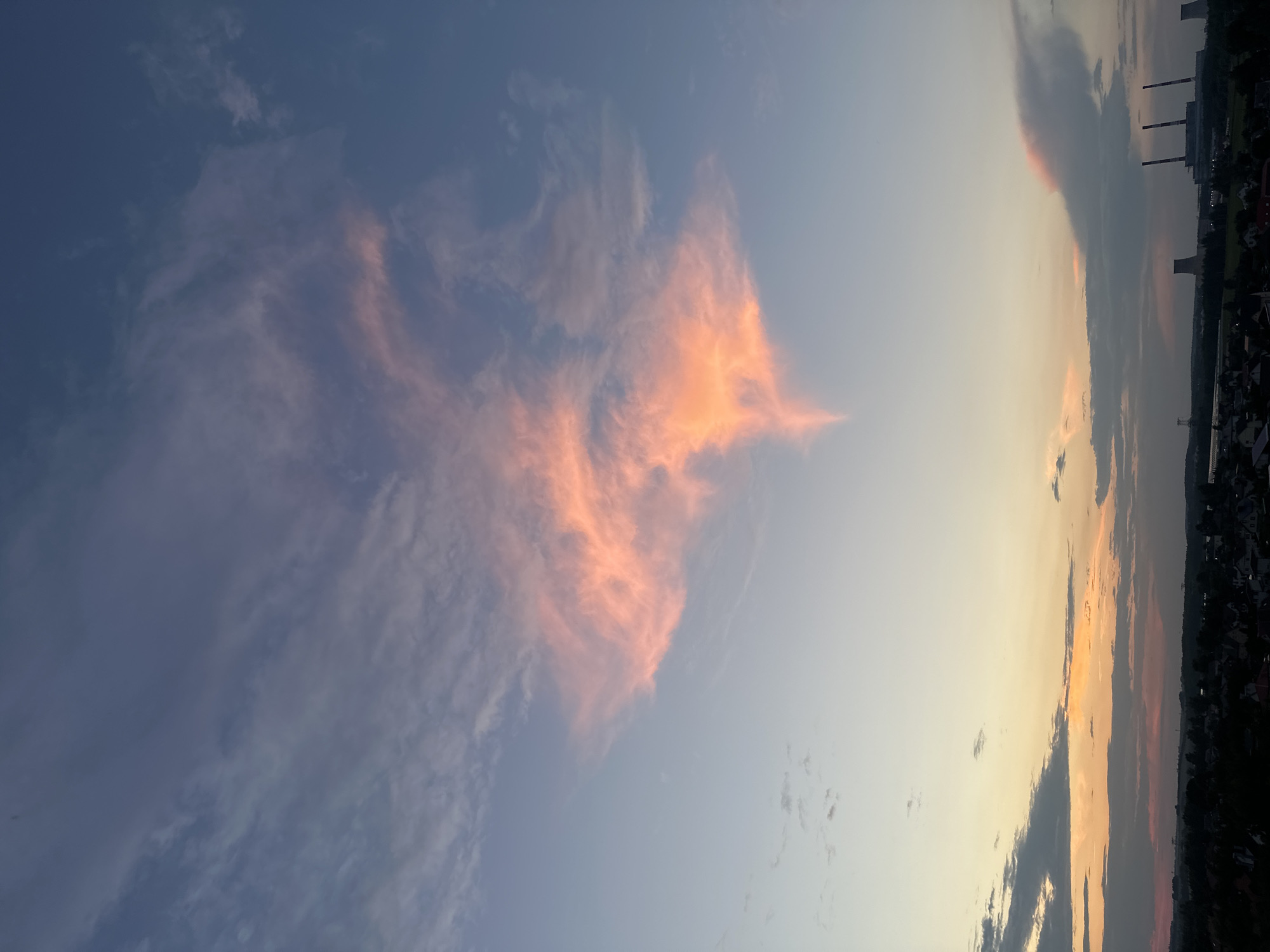 This image captures a dramatic sunset over a semi-urban landscape in the vicinity of Shchomyslitsky Sel'ski Sovet, Belarus. The sky dominates the frame, transitioning from a deep blue at the top to lighter hues of orange and yellow near the horizon. A large, wispy cloud formation in the upper center of the sky is illuminated by the setting sun, casting a vibrant pink and orange glow. This cloud is uniquely shaped, with a wider upper section tapering to a pointed tip.
Below the sky, a silhouetted horizon reveals the outlines of buildings and vegetation, suggesting a mixture of residential areas and industrial structures. In the far right of the image, three tall smokestacks belonging to a power plant are visible against the dusky sky, with a cooling tower to their left. The foreground features a dark, indistinct view of rooftops, indicating a low-lying settlement. The overall impression is one of a serene, yet industrial, twilight scene.