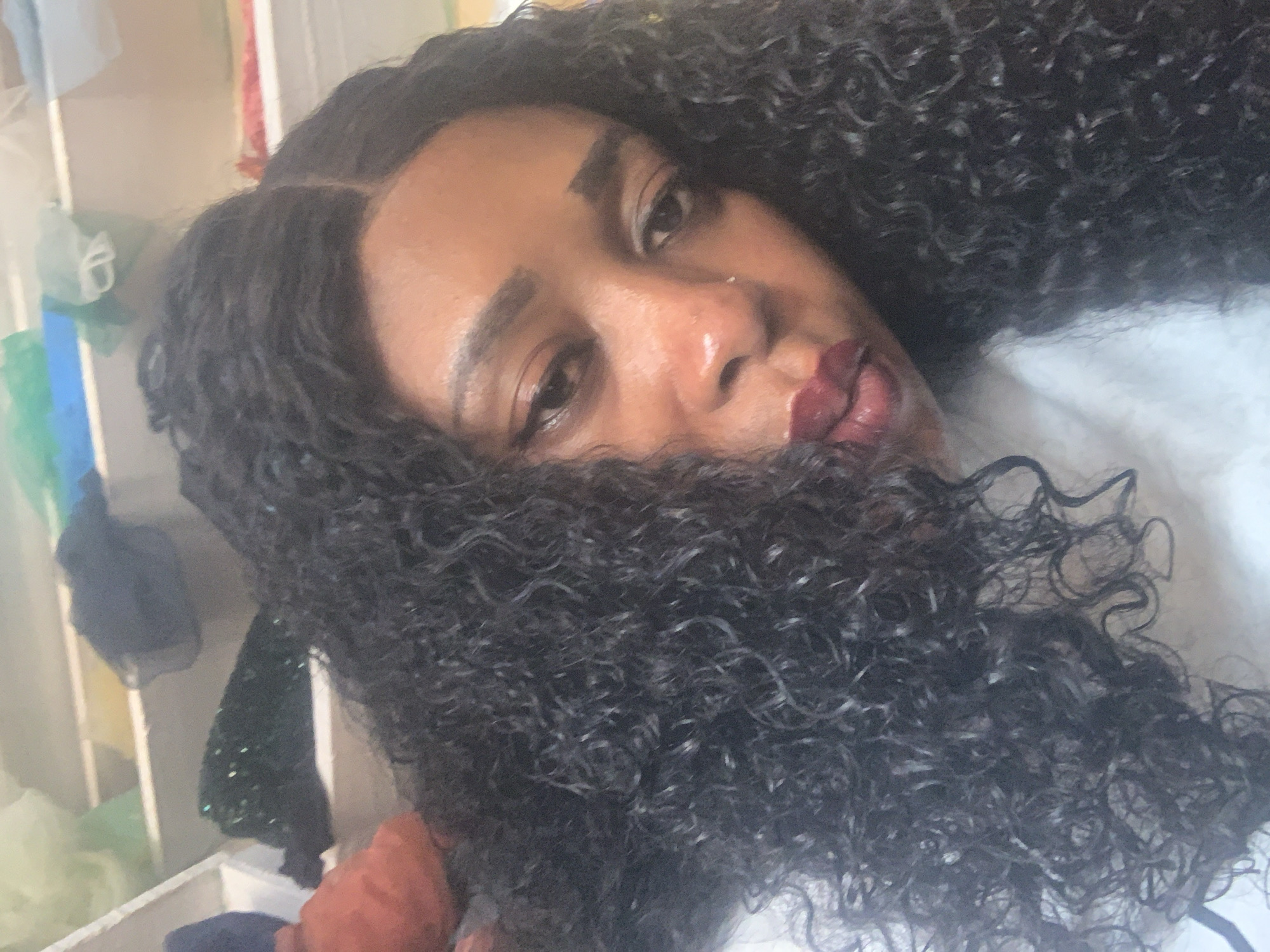 In this close-up, waist-up photograph, a Black woman with dark, curly hair and deep red lipstick is the central subject. She is positioned slightly off-center to the right, with her head tilted down and to the left, looking towards the viewer. Her eyes are partially closed, and her expression is somewhat melancholic. Her hair is voluminous and appears to be a wig, framing her face. The background is out of focus, showing shelves stacked with various items, including what appear to be fabrics or clothing in muted colors such as green, blue, and off-white. The lighting is somewhat harsh, creating highlights on her skin. The overall impression is a candid, personal moment captured within an indoor setting, possibly a home or storage area in Sabon Tasha, Nigeria.