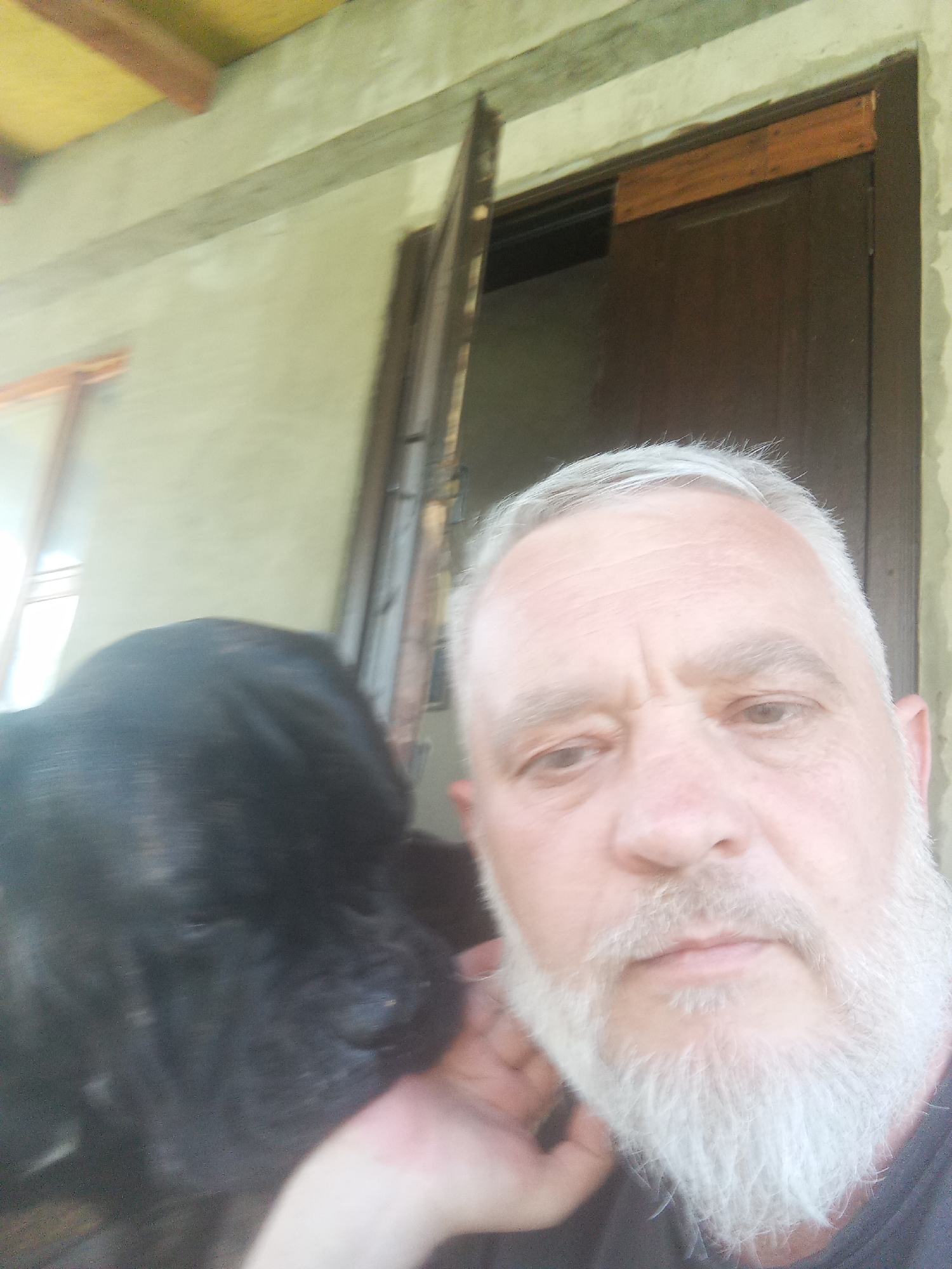 The image is a close-up, selfie-style photograph featuring a man and a black dog. The man, with a white beard and hair, is positioned on the right side of the frame, his face partially obscured by his hand and the dog. He appears to be looking towards the camera. To the left, a black dog is partially visible, with its head and part of its body in the foreground, leaning against the man.
The background suggests an outdoor setting, possibly a porch or an area with an overhang. Visible architectural elements include a light-colored wall, wooden beams, and a dark wooden door that is ajar. The lighting indicates it is daytime, with natural light illuminating the scene. The overall impression is informal, capturing a moment between a man and his pet. There are no specific landmarks or elements that pinpoint the location to the city of Brovary, Ukraine.