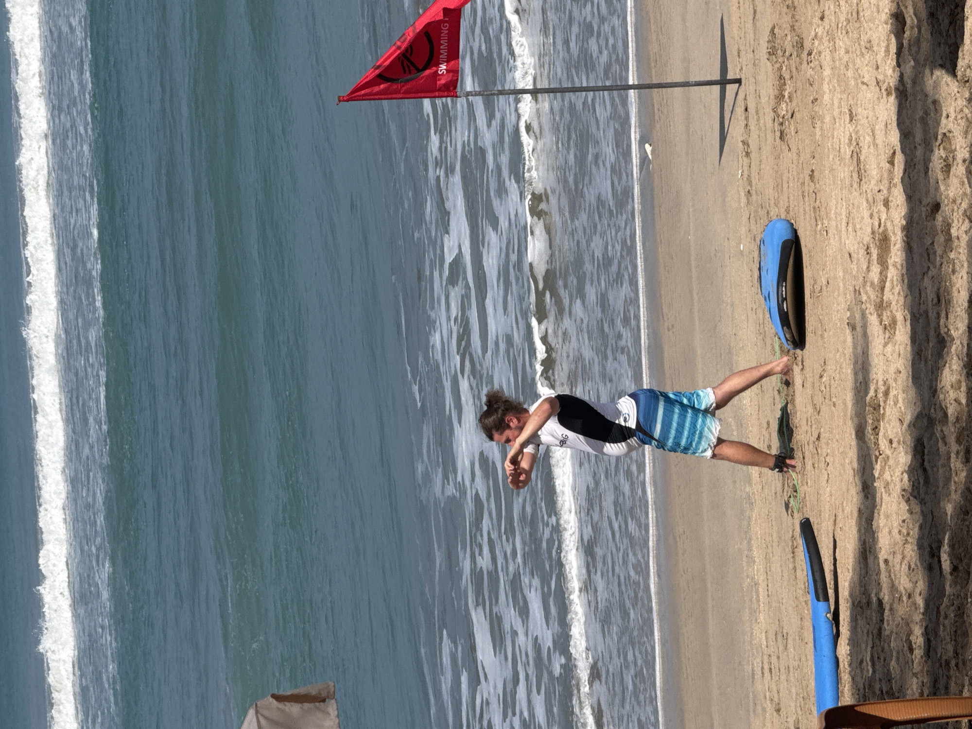 This image depicts a beach scene in Kuta, Indonesia, with the ocean and sand as the primary environments. A male individual, appearing to be engaged in a physical activity or stretching, stands on the sand near the water's edge. He is wearing a white t-shirt with black accents and blue and white striped shorts. A red flag with the word "SWIMMING" is planted on the beach, indicating a designated swimming area. Two surfboards, one blue and black, and another blue, are present on the sand, suggesting activities like surfing may be popular in this location. The ocean features moderate waves, with white foam cresting as they approach the shore. The overall atmosphere is that of a typical beach day with recreational activities in progress.