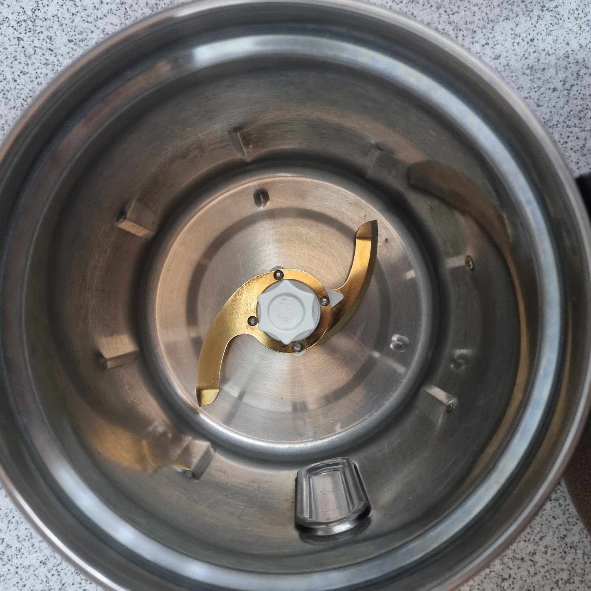A close-up, overhead shot reveals the interior of a stainless steel blender bowl. A golden-colored blade assembly with a white plastic cap at its center is positioned at the bottom of the bowl. The blender bowl is clean and appears to be a domestic appliance. The background outside the bowl shows a speckled gray surface, possibly a countertop or table. The image does not contain any information about the location in Dunnae-myeon, South Korea.