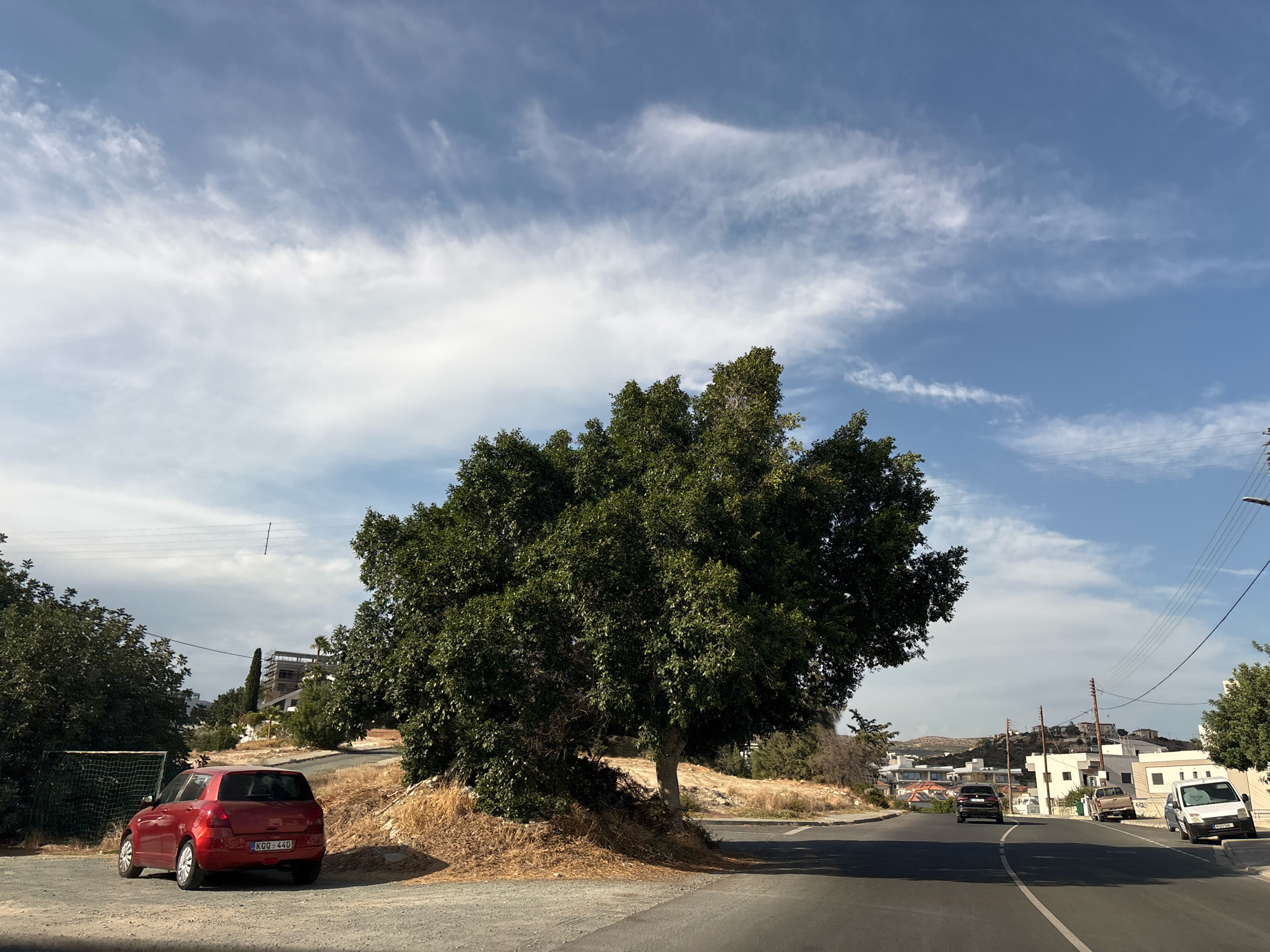 The image depicts a street scene in Agios Athanasios Municipality, Cyprus, under a partly cloudy sky. A red hatchback car, identified by its license plate KDO 440, is parked on the left side of a dirt and gravel road. Further down the road, a large, dense green tree dominates the center of the frame, with dry, straw-like vegetation at its base.
The road curves to the right, revealing a residential area with white-and-beige buildings on a hillside in the distance. Several other vehicles are visible on the road, including a black SUV, a pickup truck, and a white van. Power lines are strung across the sky, supported by poles. A soccer goal with a green net is situated behind the red car, partially obscured by foliage. The overall environment appears dry and sunny, typical of a Mediterranean climate. No people are visible in the image.