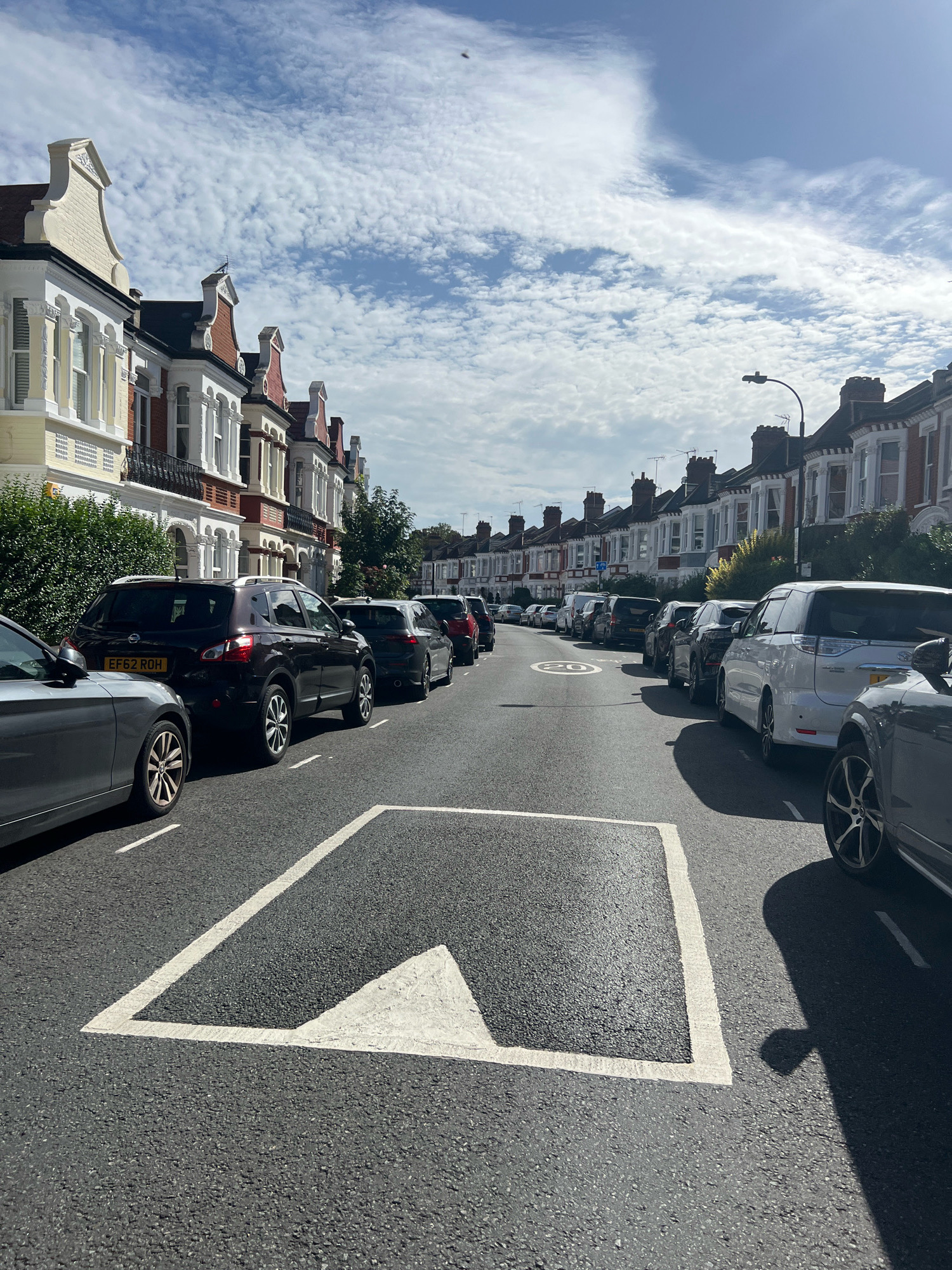 A street in London, United Kingdom is lined with terraced houses. The houses are made of brick with white and red accents. They have multiple stories and large windows. The street is paved with asphalt and has white lane markings and a speed limit sign that reads "20". Several cars are parked on the side of the street. The sky is blue with scattered white clouds.