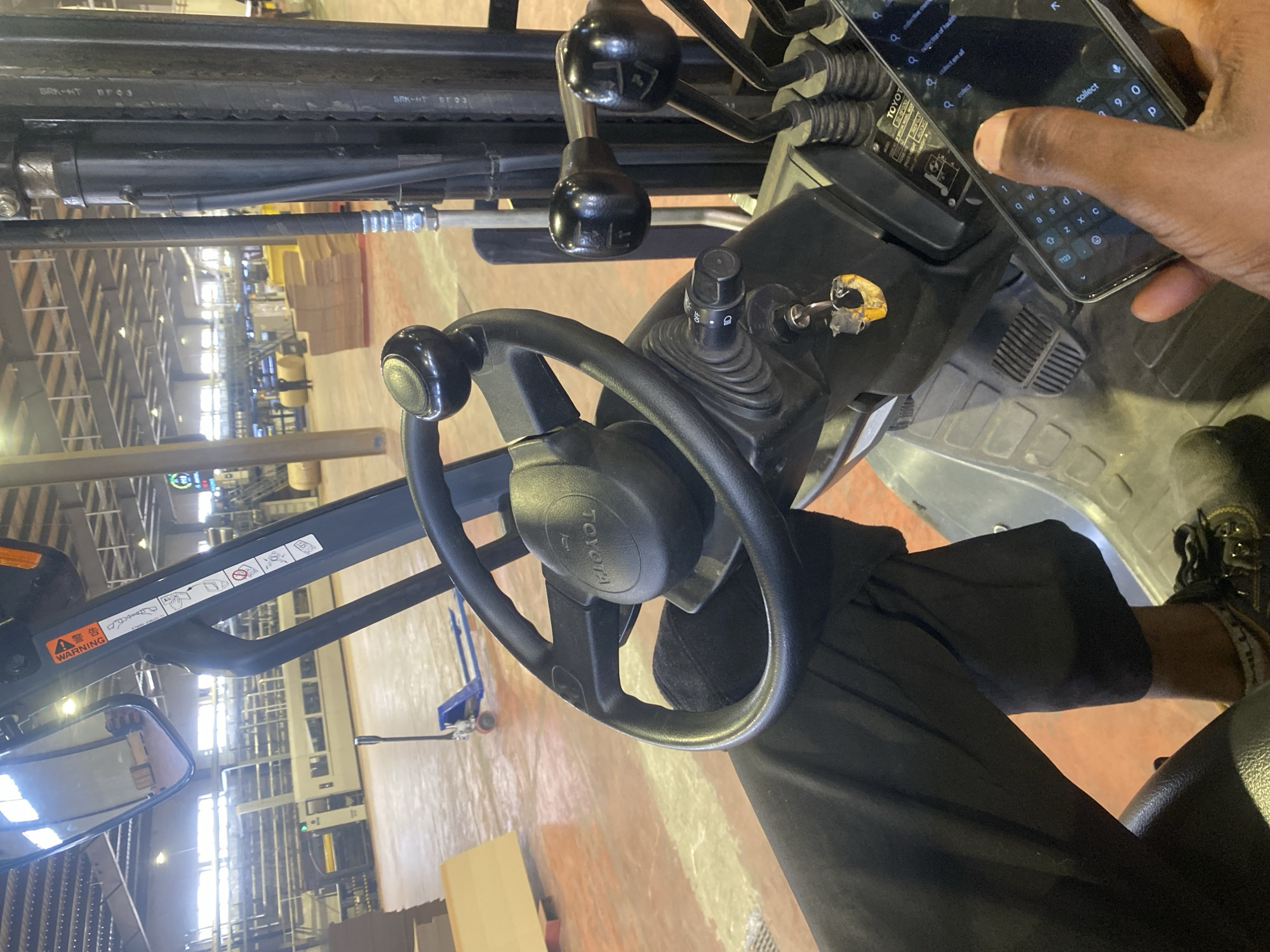 The image, captured in a portrait orientation, shows a close-up, downward-angled view from the operator's seat of a forklift within what appears to be a large industrial or factory setting in Paipe, Nigeria.
The primary subject is a person, whose right hand is actively engaged with a smartphone, typing "collect" into a search bar, while their left leg is visible near the forklift controls. The forklift itself is a Toyota model, identifiable by the "TOYOTA" logo on the steering wheel and a label near the phone. Other visible parts of the forklift include various control levers, an ignition key in place, a switch marked "OFF," and safety warning labels on the overhead guard that read "WARNING" along with graphic safety symbols. The front portion of the forklift's frame and hydraulic lines are visible, with text "SRK-HT" and "BF03" on parts of the machinery. A side mirror reflects part of the factory interior.
In the background, the factory floor stretches out, showing industrial lighting, structural beams, and what looks like other machinery or stacked goods. A blue hand cart is also visible on the floor. The activity clearly shows the person operating a smartphone while seated at the controls of a powered industrial truck, indicating a potential distraction during operation. The setting suggests daytime due to the bright ambient light within the facility.
Visible text includes: "WARNING," "SRK-HT," "BF03," "TOYOTA," "OFF," "collect," "collect em all," "collective me...," and numerical/alphabetical characters from the phone's keyboard like "7123," "0," "p," "b," "c." A label also displays "32-07-030."