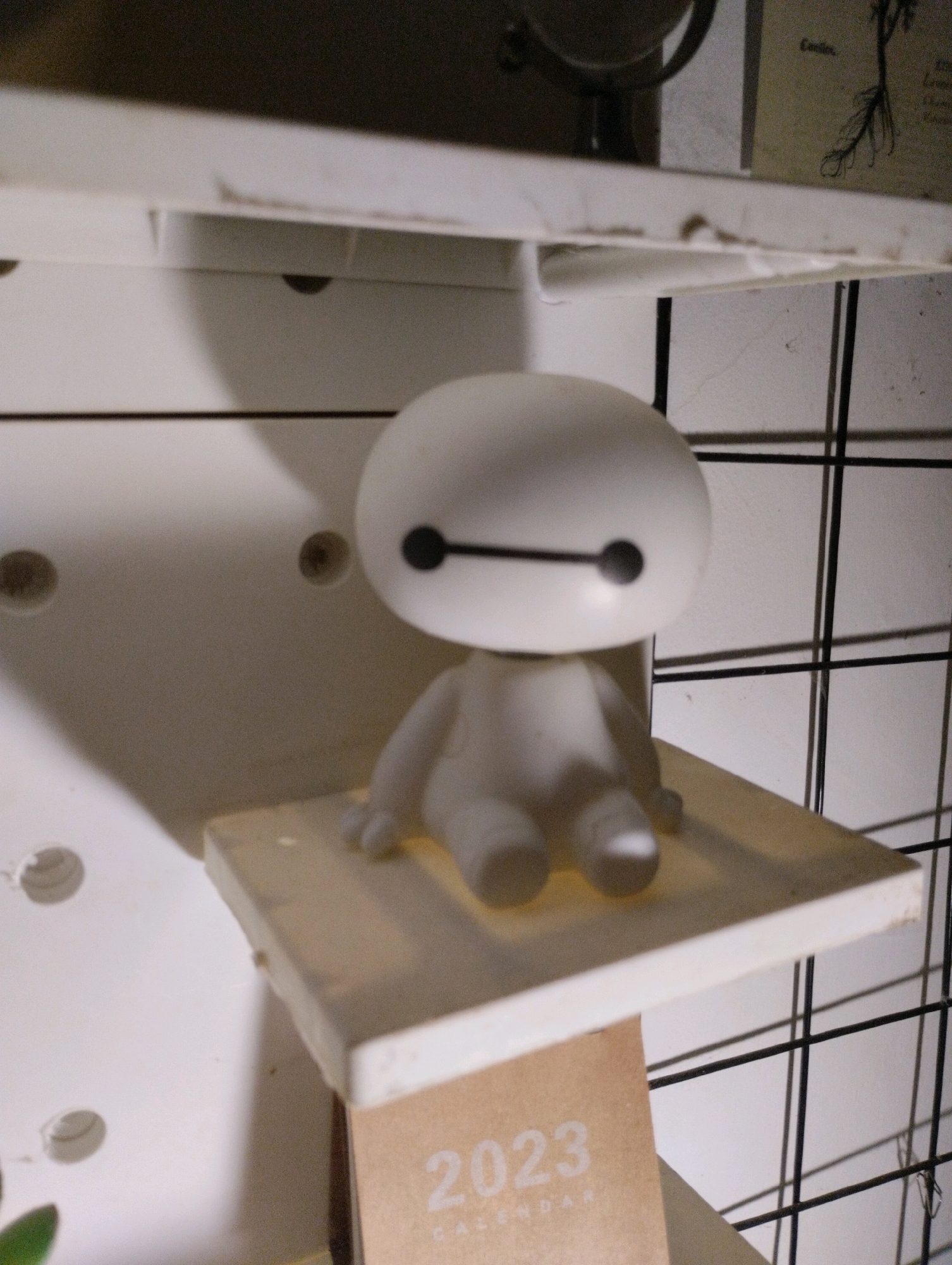 A white figurine resembling Baymax is sitting on a small, light brown shelf. The figurine has a round head with two black dots for eyes connected by a horizontal black line. Below the head, it has a simple white body with stubby limbs. The shelf is placed on a white pegboard with several circular holes visible. To the right of the figurine, a black grid shelf is present. In the foreground, a portion of a brown calendar with the text "2023 CALENDAR" is visible. The scene appears to be indoors, possibly in a room within the city of Cimenyan, Indonesia, given the specific location prompt. The lighting suggests a dimly lit environment with shadows cast from the figurine and the shelf onto the pegboard.