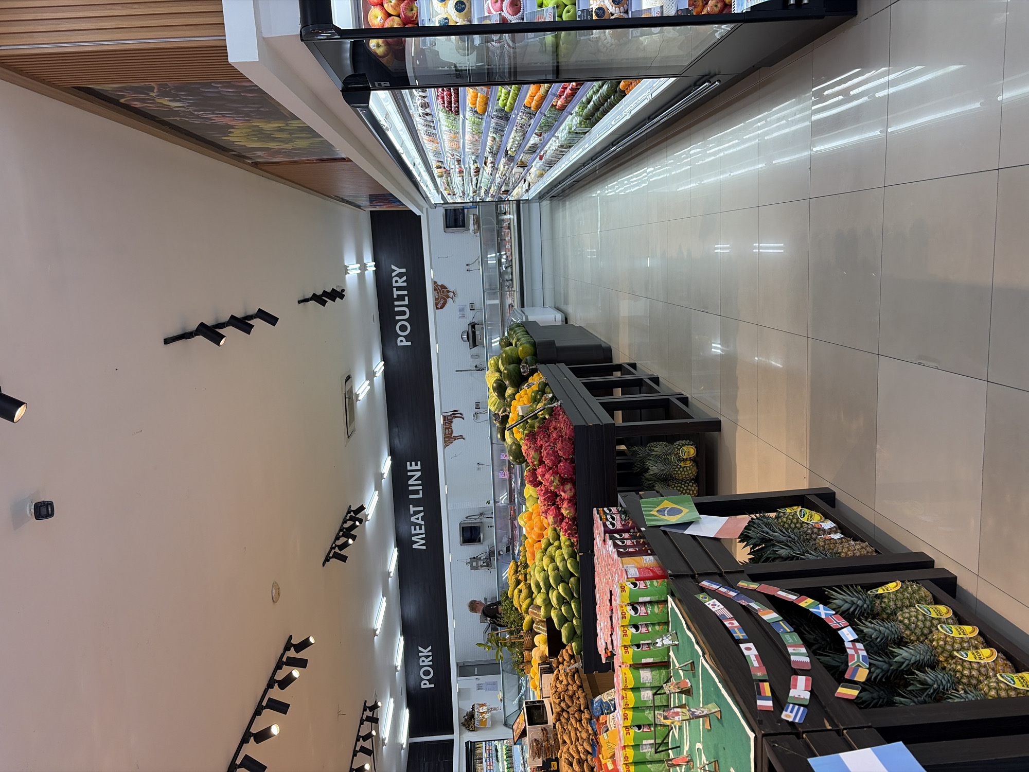 The image depicts the interior of a brightly lit supermarket in North Kuta, Indonesia, with a focus on the produce and meat sections. In the foreground, various fruits are displayed on tiered wooden shelves, including large piles of sweet potatoes, watermelons, mangoes, and dragon fruit. Several cans of Pringles chips are also visible on a shelf. Decorative strings of international flags hang above the fruit displays, suggesting a possible event or promotion. Further back, the "MEAT LINE" and "POULTRY" sections are clearly marked with black signage on a white wall, with refrigerated display cases showing various meat products. A man, presumably a store employee, is visible in the background near the meat section. To the right, a long aisle is lined with illuminated refrigerated shelves stocked with a wide assortment of fruits and vegetables. The floor is tiled and reflects the overhead lighting.