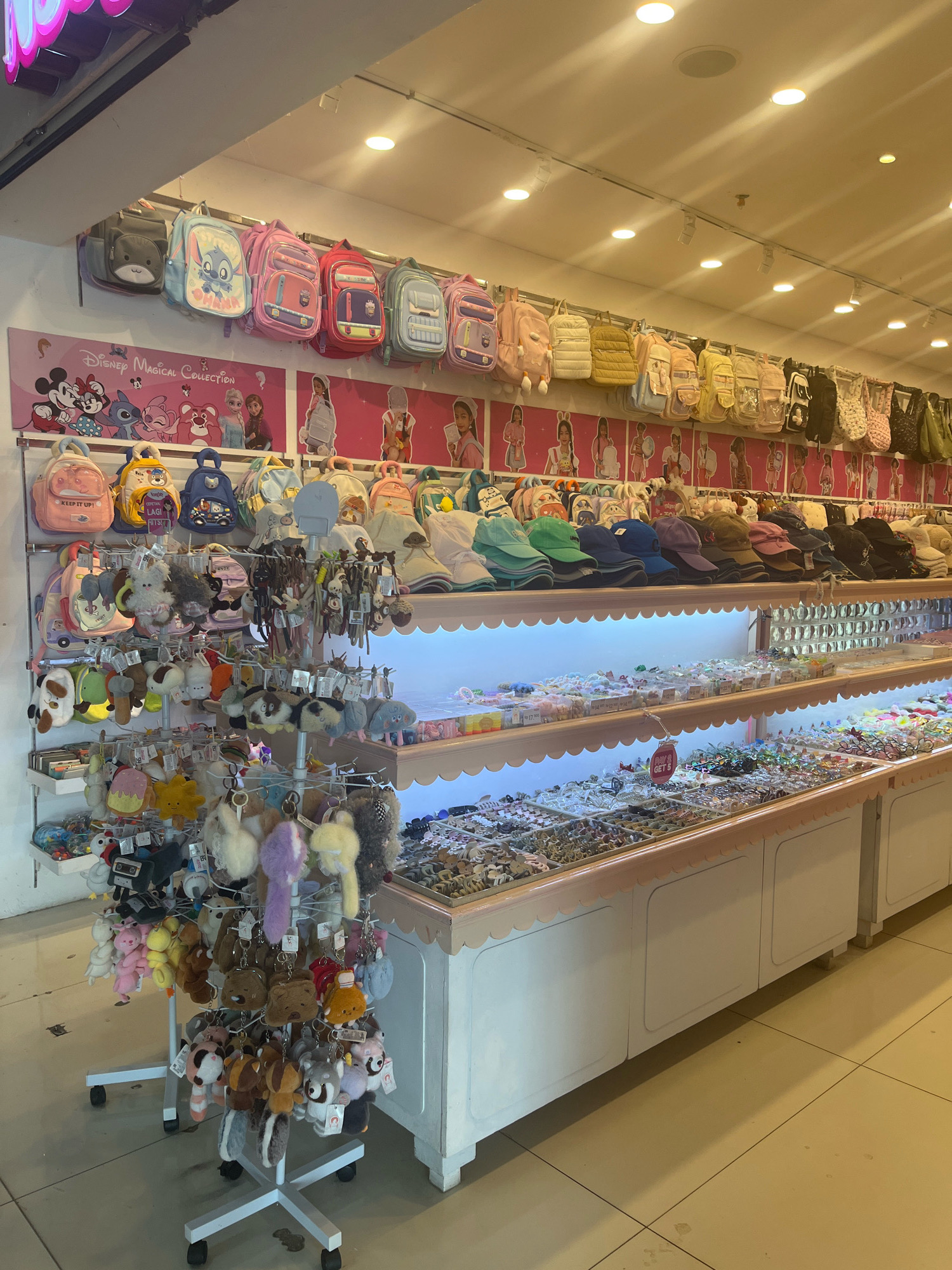 The image shows a retail store that appears to sell children's accessories and school supplies. The store is brightly lit with overhead lighting and displays a wide variety of products on shelves and hanging racks.
In the upper section of the image, several colorful backpacks are displayed, many featuring cartoon characters like Disney's Mickey Mouse and Stitch. Below these, shelves are filled with smaller bags, pouches, and plush keychains in various animal and character shapes. Further down, rows of hair accessories such as clips, ties, and headbands are neatly organized in clear display cases. The overall aesthetic is playful and geared towards a young audience. The store has a clean, organized layout with light-colored walls and shelving. The floor is tiled. The image does not contain any people. The store appears to be located in Sengkang, Indonesia.