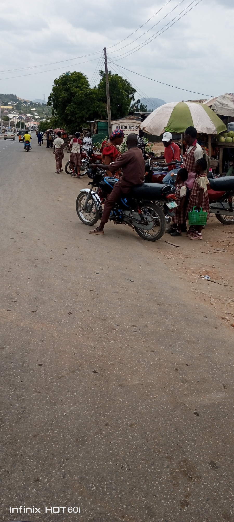 The image shows a street scene in Zango-ndaji, Nigeria. A man sits on a black and silver motorcycle parked on the side of the road. Two young girls in school uniforms stand beside the motorcycle, one holding a green basket. Other people stand nearby, including some under a white and green striped umbrella. Several motorcycles are parked along the side of the road. A sign in the background reads "Welcome to the Palace of HRH (ELDER) BAYO JOH". The road surface is grey asphalt. Buildings and hills can be seen in the distance. The sky is overcast.