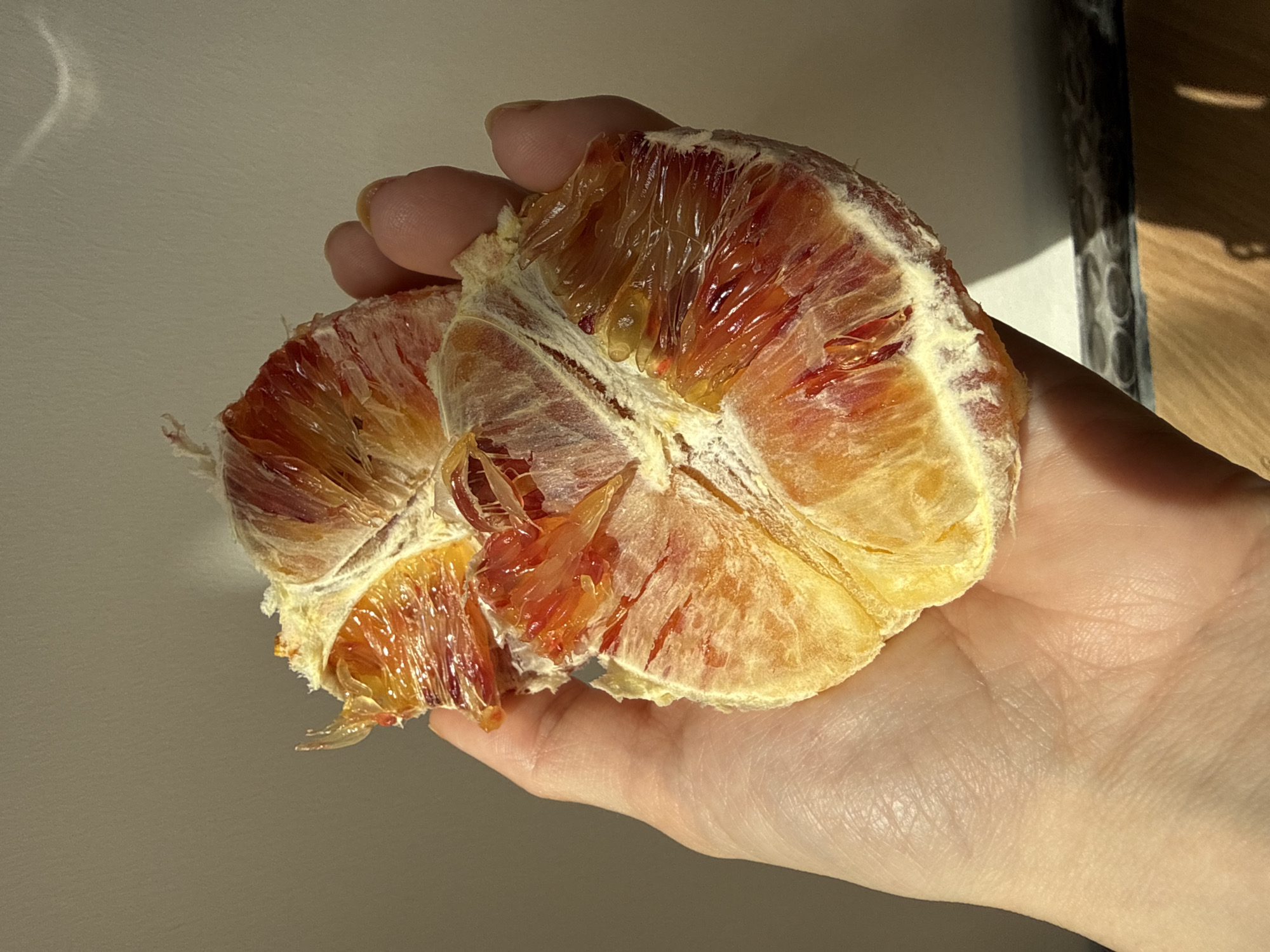 A partially peeled blood orange sits in the palm of a hand. The orange is segmented, showing red and yellow flesh. The hand is fair-skinned. The background is a gradient of light to dark, suggesting a room interior. No information indicates a location in Moscow, Russia.
