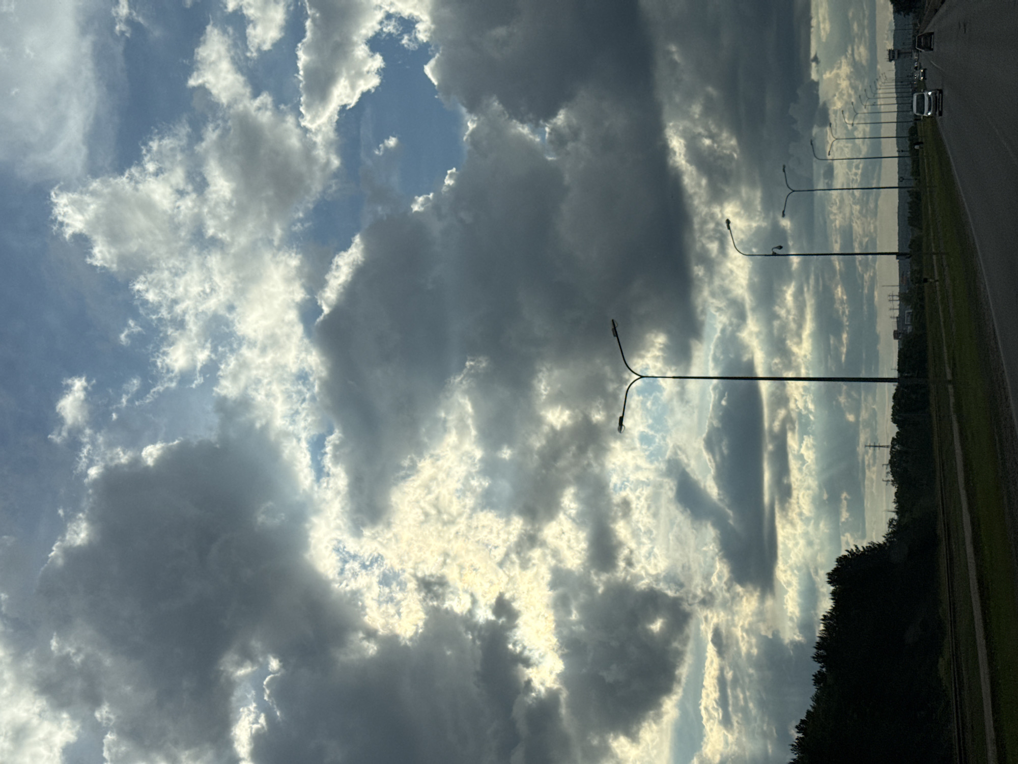 The image depicts a cloudy sky above a road in Chaikovsky, Russia. The sky features a mix of light and dark clouds, with patches of blue visible. A line of streetlights stretches along the right side of the road, and several cars are visible in the distance. A line of trees and shrubbery is visible along the right edge of the frame. The image is oriented vertically.