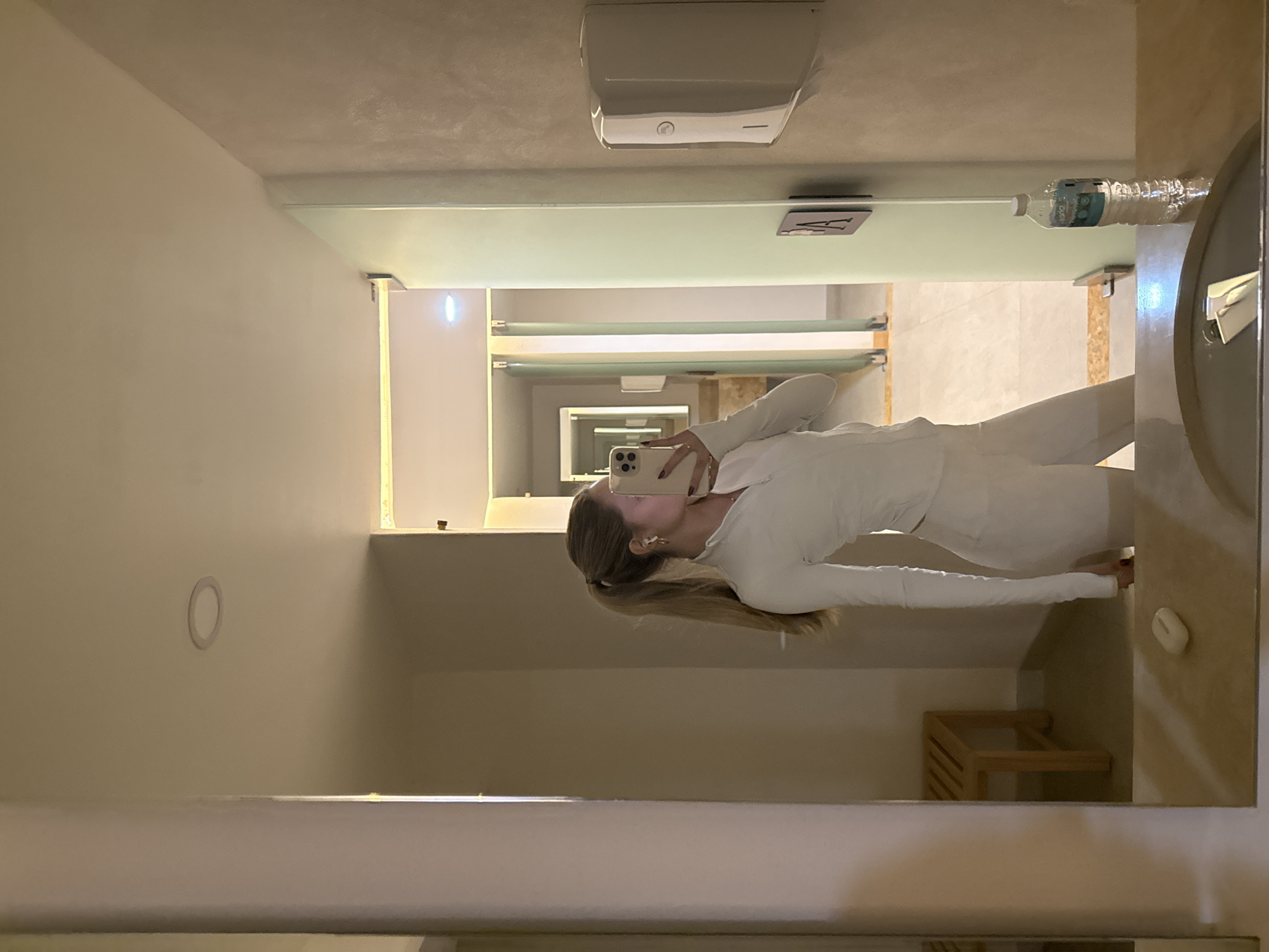 The image is a mirror selfie taken indoors, depicting a female subject in what appears to be a public restroom or changing facility in Mexico City, Mexico.
The subject is a light-skinned female with blonde hair tied in a high ponytail. She is wearing a form-fitting white athletic ensemble, consisting of a long-sleeved, quarter-zip top and matching shorts or leggings. She has white wireless earbuds in both ears and a small gold hoop earring in her visible left ear. Her face is mostly obscured by a light-colored smartphone, held in her right hand, which she is using to capture her reflection. Her left hand, with dark nail polish, is also visible.
On the light-colored countertop in the foreground, a white wireless earbud charging case is positioned to the left of the subject's reflection. To the right, a clear plastic bottle of Jarritos-branded water stands upright. A metallic sink faucet is partially visible within the basin.
The environment features light-colored walls and ceiling, with circular recessed lighting fixtures visible. To the left, a slatted wooden bench is partially reflected. To the right, a white wall-mounted dispenser (likely soap or sanitizer) is visible above a dark square sign attached to a light green partition, displaying a partial 'PA' or 'BA' character. Beyond the subject, a series of individual stalls or changing rooms with frosted or light-colored glass doors and vertical metal frames extend down a brightly lit hallway. The flooring consists of light-colored tiles, some with darker contrasting strips. The presence of a Jarritos-branded water bottle indicates a Mexican location.