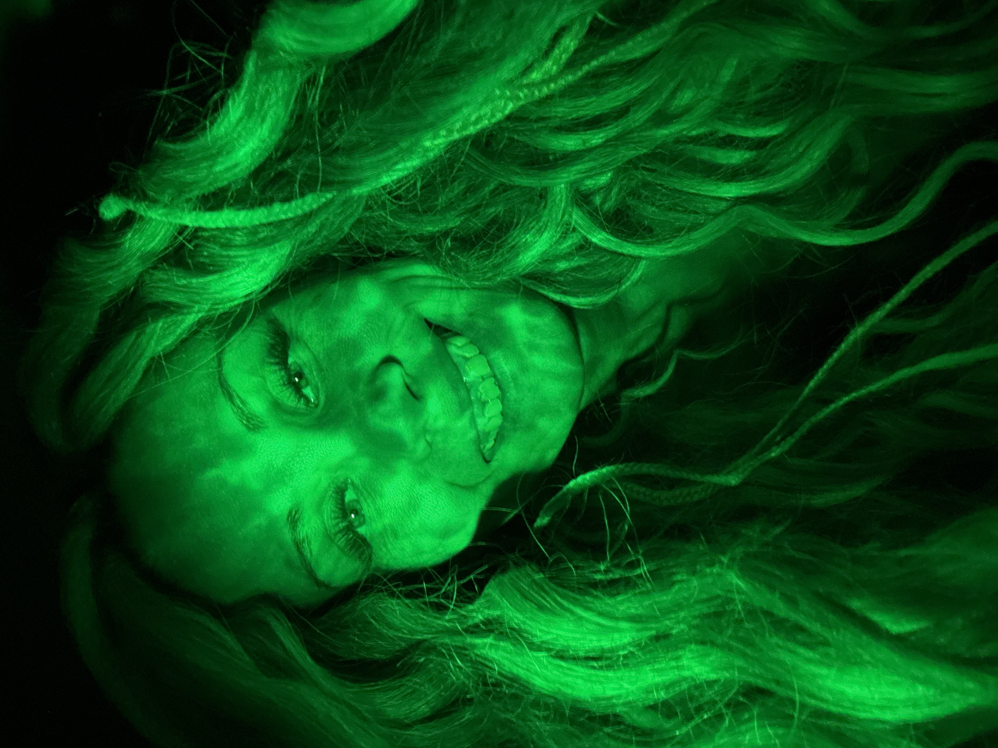 A close-up, vertically-oriented image shows a woman's face amidst a mass of long, wavy hair. The entire image is rendered in shades of green. The woman's head is tilted slightly, with a partial smile revealing her teeth. Her eyes are open, gazing upwards. The surrounding hair partially obscures her face. The background is a stark, dark green or black, contrasting with the brighter, more illuminated green of the subject. The image lacks specific identifiable features linking it to Rusanovo, Russia.