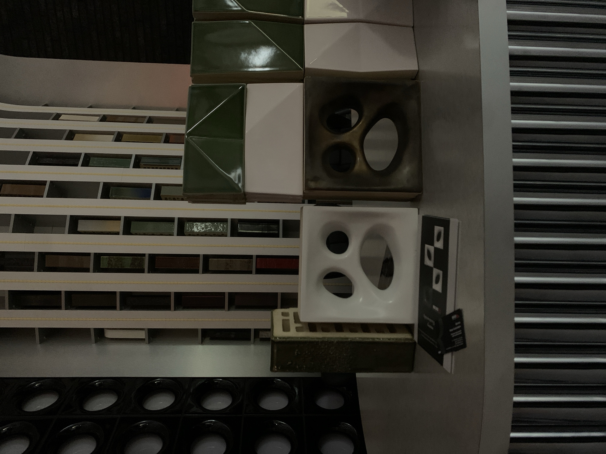 The image shows a close-up of various tile samples in a store, possibly located in Moscow, Russia. The samples are displayed on shelves, with some stacked on top of each other. The tiles vary in color, shape, and texture, with some featuring geometric patterns and others with abstract designs. The background includes a row of recessed lights and a shuttered wall. The scene appears to be indoors.