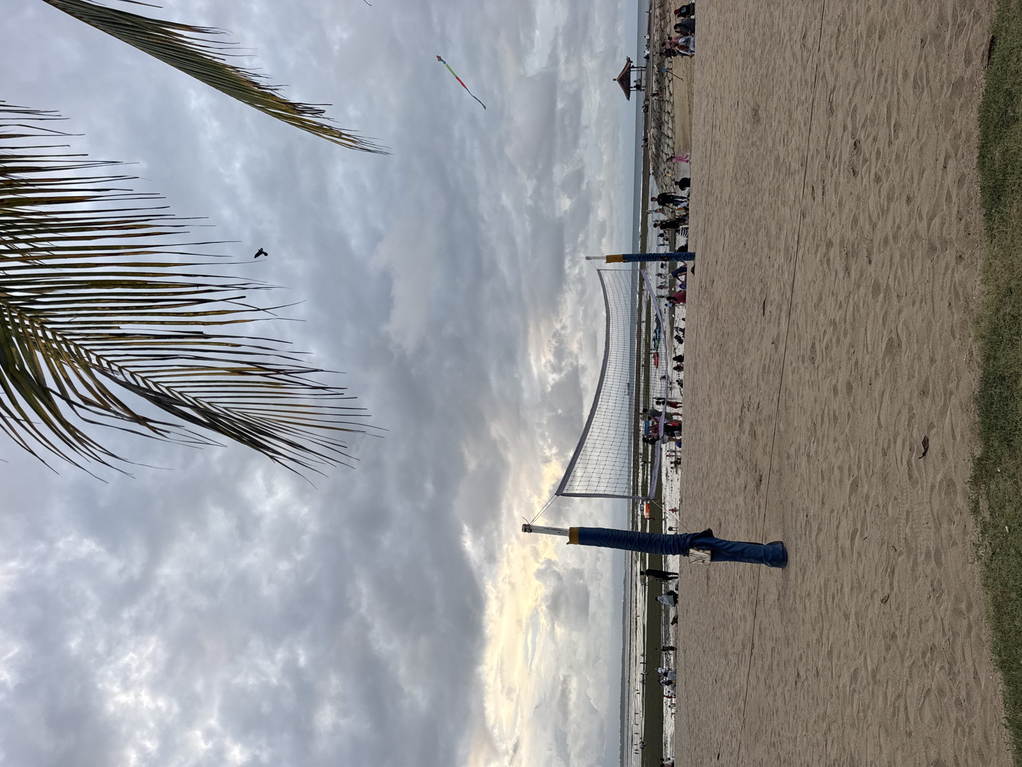 The image depicts a beach volleyball court on the beach of Kuta Selatan, Indonesia. The volleyball net is visible, with blue and yellow poles. The beach has multiple people on it. A palm tree is on the left side of the image, and grass is on the right side. There is a kite in the sky. The sky is cloudy.