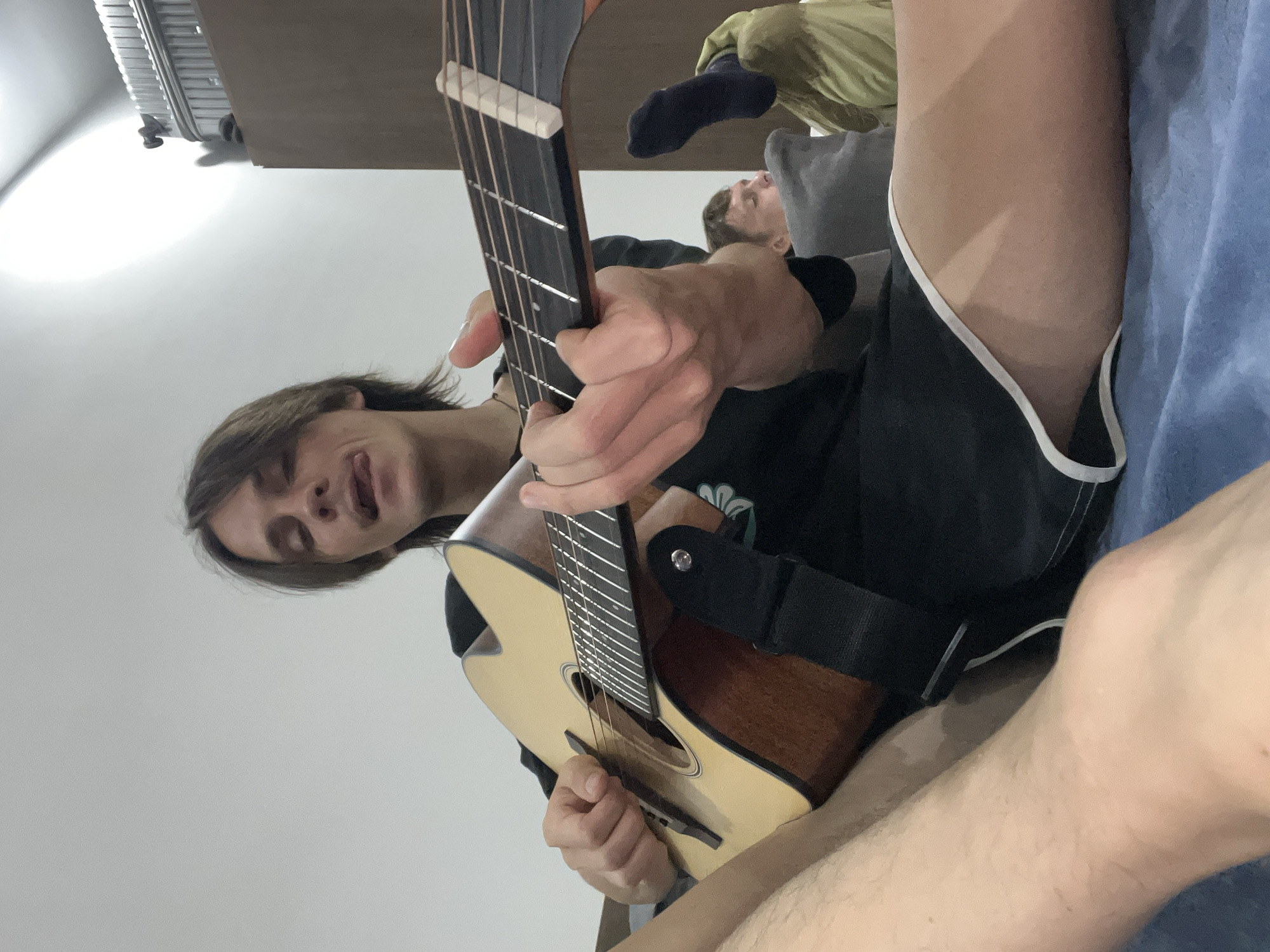 The image shows a young man playing an acoustic guitar indoors. He is sitting on a blue surface, possibly a bed or sofa. His tongue is sticking out, and he appears to be focused on playing. Another person is visible in the background, partially obscured. The room is lit by a bright light source overhead. There is a piece of luggage on a shelf in the background. The scene is not specific to any particular location in the city of Đà Nẵng, Vietnam.