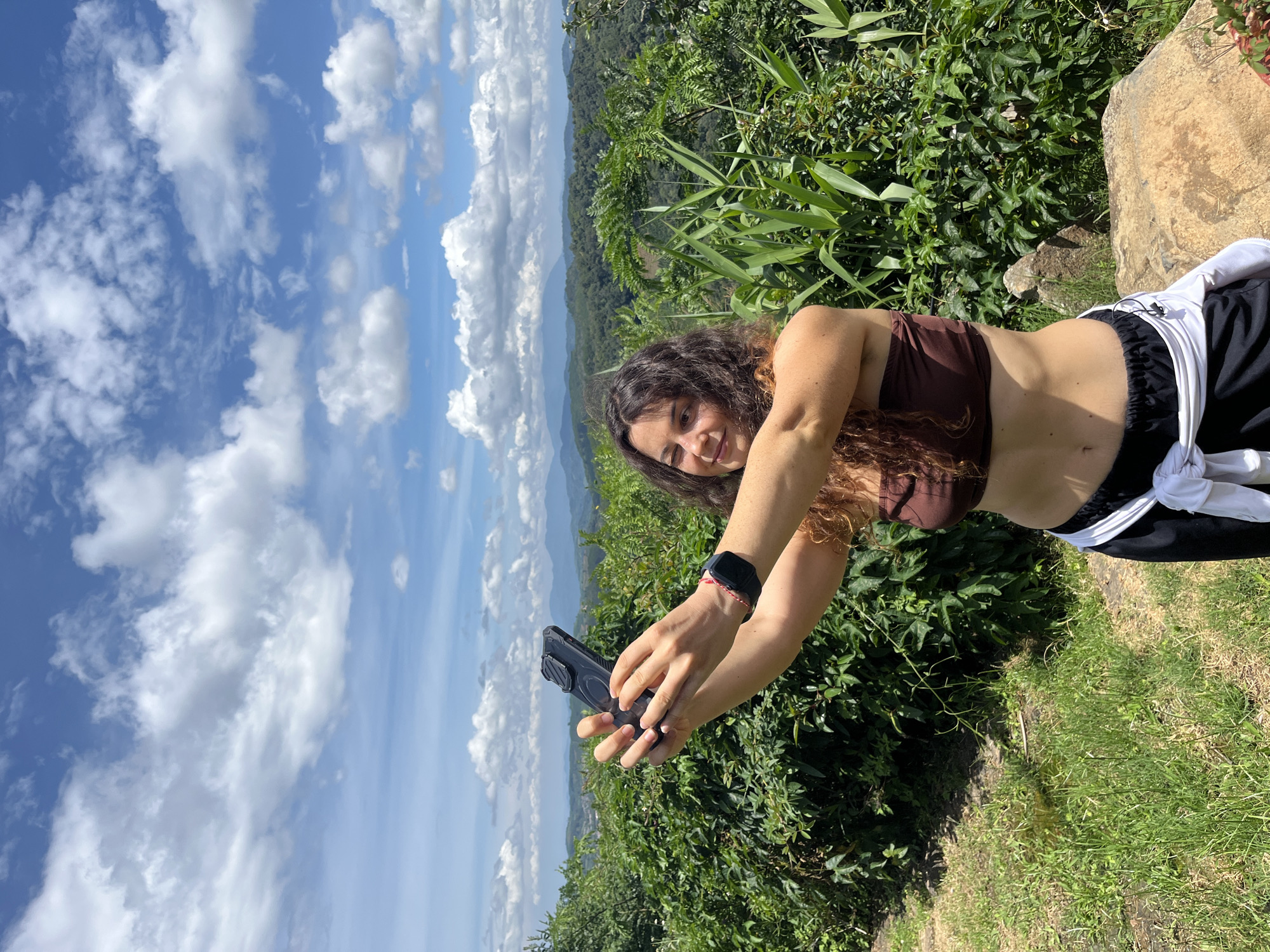 In Sơn Điền, Vietnam, a woman with curly brown hair, wearing a brown bandeau top and black pants with a white shirt tied around her waist, stands outdoors, taking a selfie with her phone. She is positioned in the foreground, with a lush green hillside covered in dense vegetation and trees behind her. The background reveals rolling hills and mountains under a bright blue sky filled with fluffy white clouds. A large rock is visible to her right. The overall scene depicts a scenic natural environment, likely a viewpoint or hiking trail.