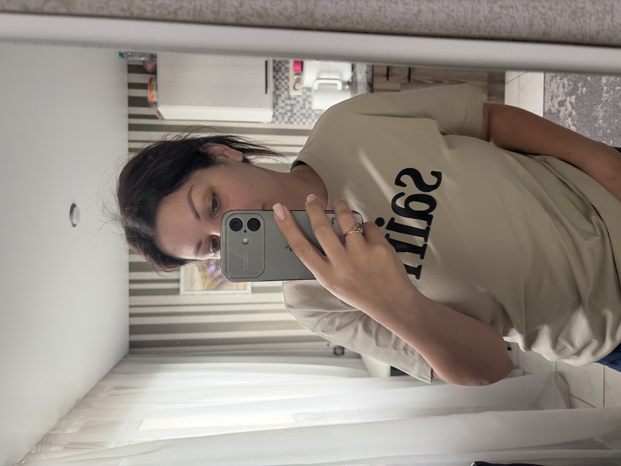 A woman with dark hair is taking a mirror selfie indoors. She is wearing a light-colored t-shirt with black lettering that reads "saint62". She has a ring on her left ring finger and her nails are painted. The background appears to be a room with striped wallpaper and white curtains. Kitchen cabinets and appliances are visible in the reflection of the mirror. This image was taken in Sochi, Russia.