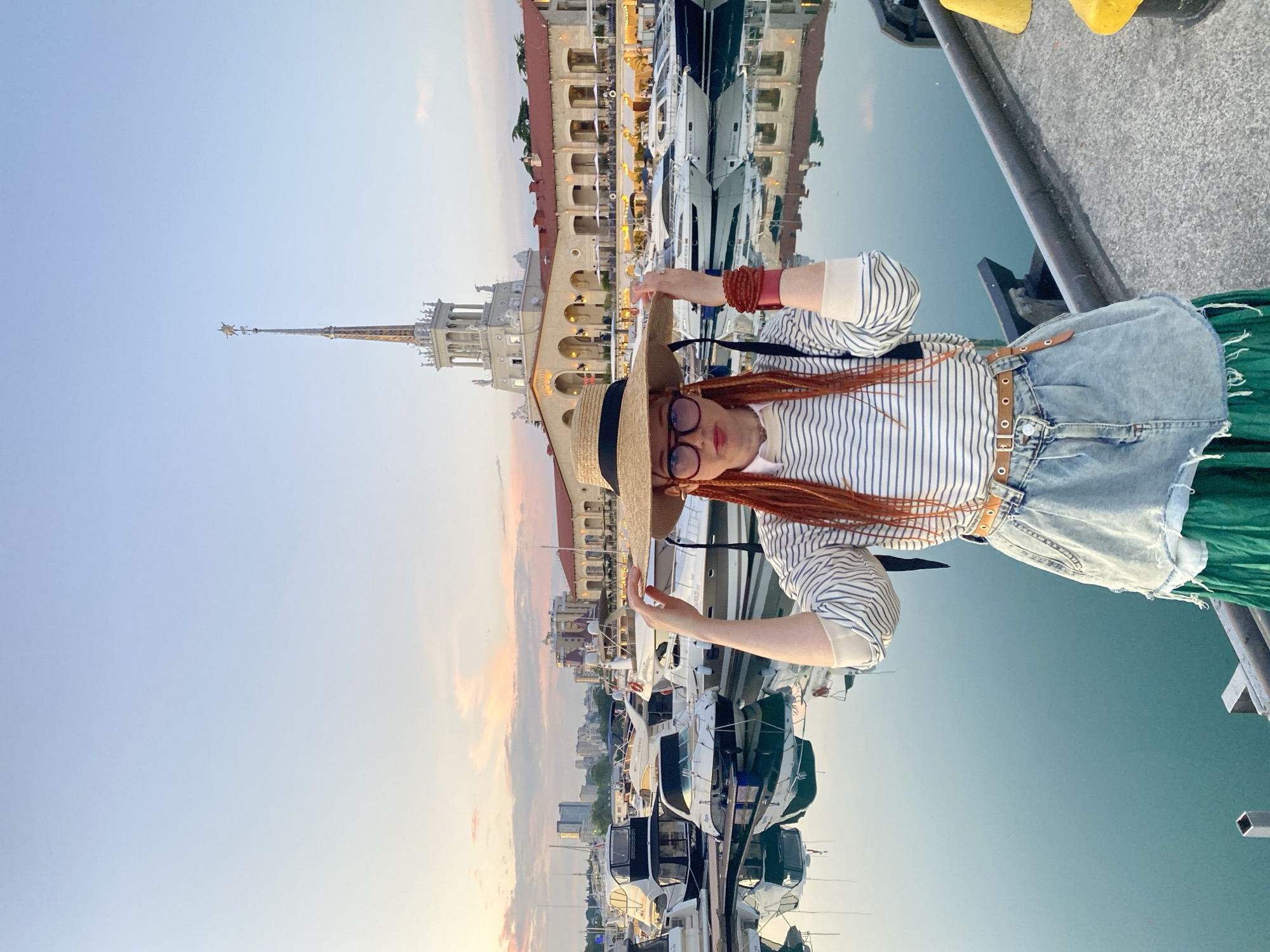 A person with ginger braids and a straw hat stands on a pier at the Sochi Sea Port in Russia. The individual wears a striped long-sleeved shirt, a denim skirt, and a green skirt. They are holding the brim of their hat with both hands. In the background, a row of yachts is docked on the water. Behind the yachts, a large, white building with a tall spire stands prominently. The sky is clear and pale blue, with a hint of pink and orange from the sunset. The water reflects the boats and the building.