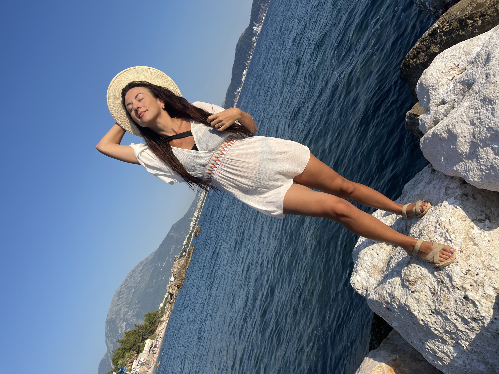 A woman stands on large white rocks near the sea in Bar, Montenegro. She is wearing a white cover-up, a straw hat, and sandals, with her eyes closed and her hand raised behind her head. The sea is a deep blue, and in the background, a coastline with buildings and mountains is visible under a clear sky. The image appears to be taken on a sunny day.