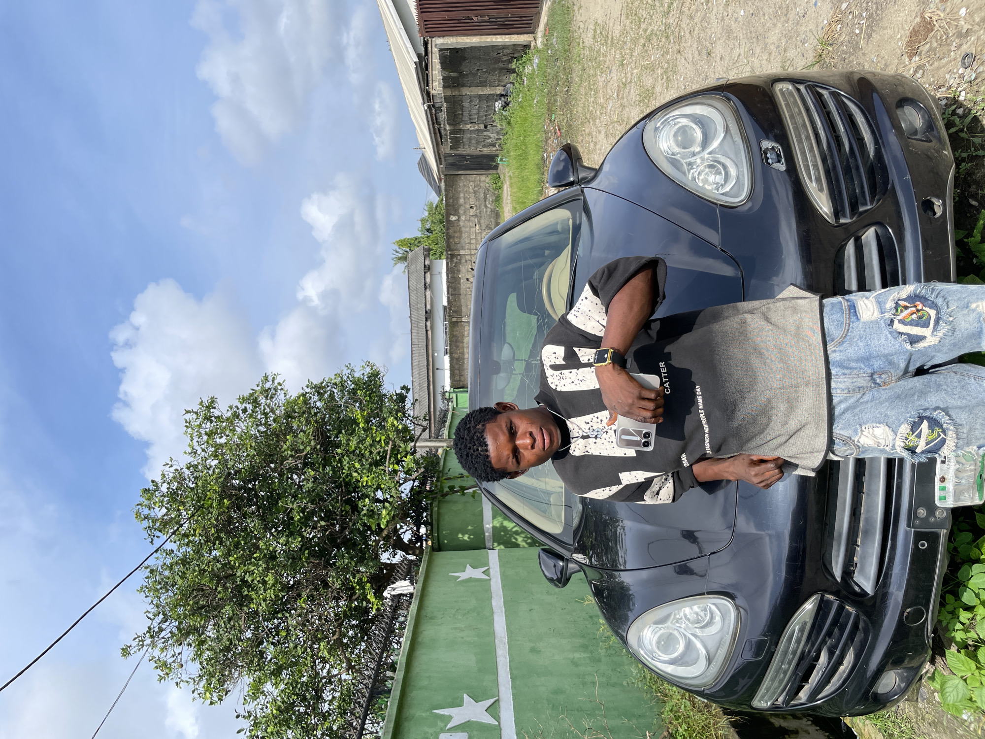 The image depicts a young male subject posing in front of a black Porsche Cayenne SUV on an unpaved residential street in Ado, Nigeria.
The sky is bright blue with scattered white cumulus clouds. Two dark utility lines traverse the upper portion of the frame diagonally. To the left, a vibrant green concrete wall features two white five-pointed star emblems. Behind this wall, a large, leafy green tree is visible, partially obscured by a dark metal fence below it. On the right side of the background, multiple low-rise structures with corrugated metal roofs are visible, separated by unpainted concrete block walls and an unkempt patch of dry grass and dirt. A reddish-brown metal gate is visible on the far right.
A black, first-generation Porsche Cayenne SUV (likely a 957 model) is parked centrally, facing the viewer. The vehicle's front bumper exhibits signs of wear, including a missing circular cover on the right side and a potential missing tow hook cover on the left. The headlights are dual projector-style units. The beige interior is partially visible through the windshield.
The male subject, appearing to be of African descent, stands directly in front of the SUV, leaning his left arm on the vehicle's hood. He looks directly at the camera with a neutral to slightly serious expression. He has short, dark, curly hair. He is wearing a long-sleeved black shirt with white and gray abstract patterns. His light-blue denim jeans are distressed with ripped sections and visible patches on the knees. On his left wrist, he wears a gold-toned wristwatch with a black strap. In his right hand, held casually by his side, he holds a light-colored smartphone equipped with a dual-lens camera module. A silver-colored pendant hangs from a necklace around his neck.
The ground in the foreground consists of compacted dirt mixed with sparse green vegetation and scattered debris.