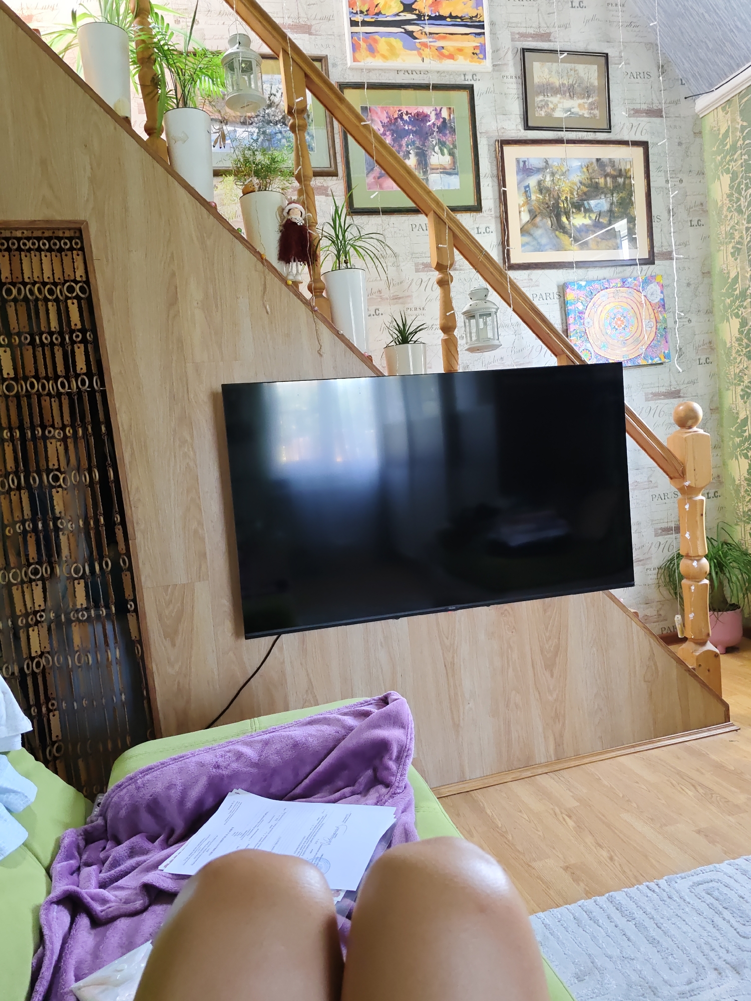 The image depicts an indoor scene, likely a living room area featuring a staircase with wooden railings and steps. Several potted plants are placed on the staircase, contributing to a sense of home decor. Framed pictures hang on the wallpapered wall beside the staircase, adding artistic elements to the space. A large, flat-screen television is mounted on the wall, positioned centrally in the frame. To the left of the staircase, a decorative beaded curtain hangs, adding a unique textural element to the scene. In the foreground, a person's legs are visible, resting on a green couch covered with a purple blanket. Papers are laid out on the blanket, suggesting a relaxed or work-related activity. The flooring appears to be made of light-colored wood. The overall impression is one of a cozy, lived-in domestic environment. There is no specific information in the image that would allow for identification of its location in Rostov-on-Don, Russia.