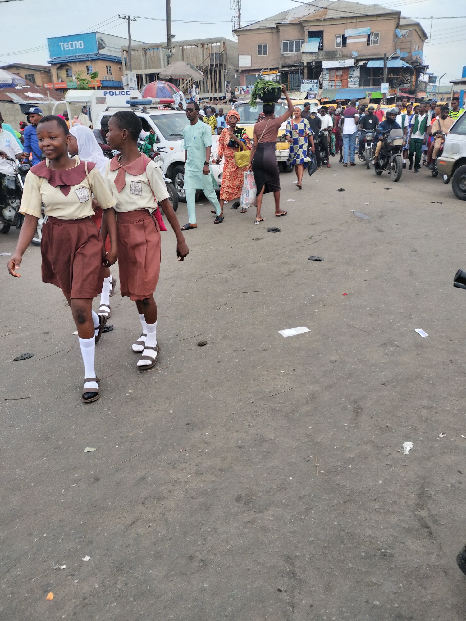 The image captures a busy street in Lagos, Nigeria. Two young girls in school uniforms, consisting of tan shirts with brown collars and brown skirts, are walking on the left side of the street. They are wearing white socks and sandals. Behind them, a white police vehicle is visible, along with numerous people walking and riding motorcycles. Buildings and businesses line the street, including a store with the sign "TECNO" on the roof. The road surface is asphalt and littered with debris. People are engaged in various activities like walking, carrying items on their heads, and riding motorcycles. The scene suggests a bustling urban environment with a mix of pedestrians, vehicles, and commercial activity.