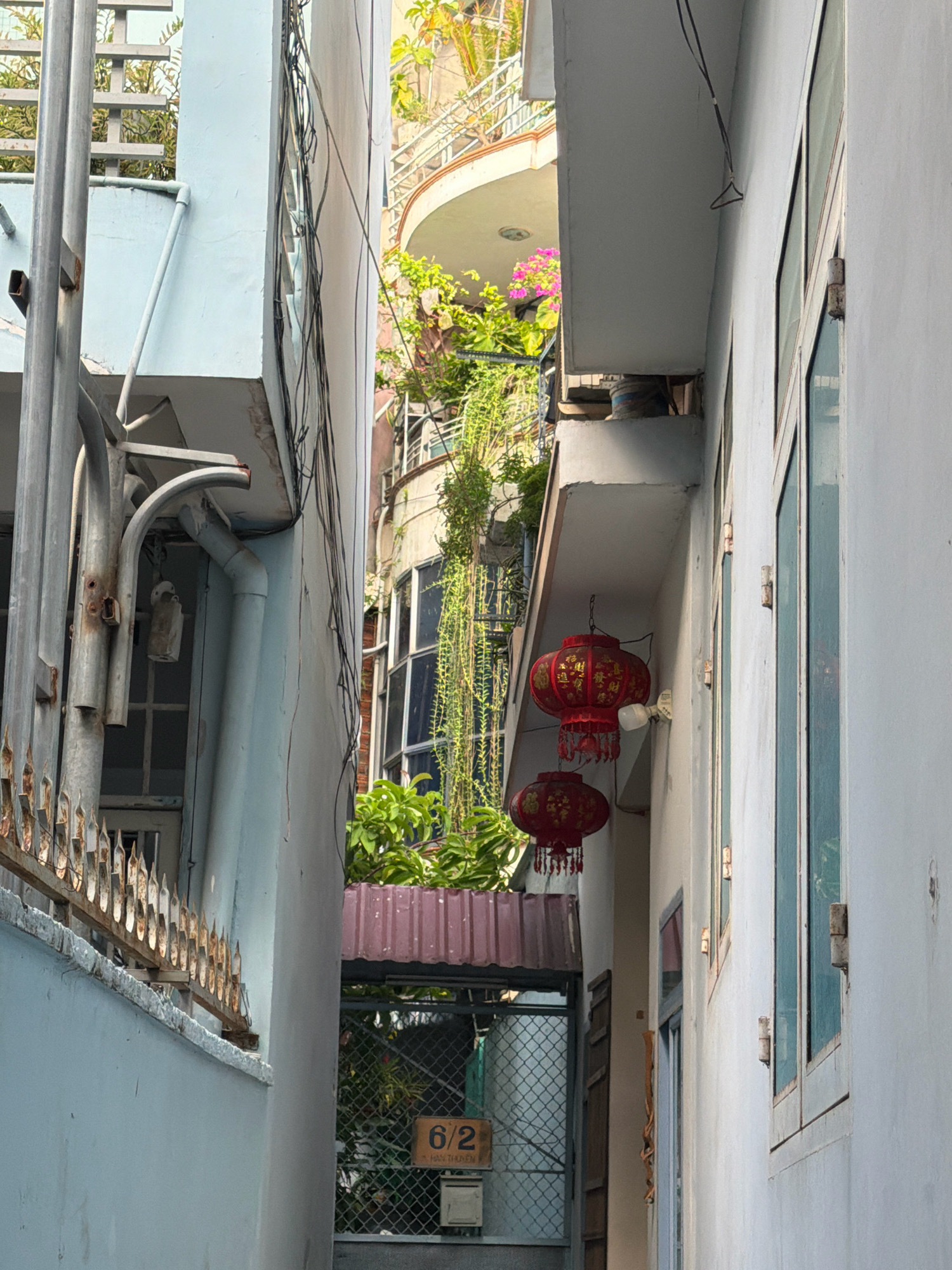 This is a photograph of a narrow alleyway between two buildings in Nha Trang, Vietnam. The buildings are white and light blue, with peeling paint and exposed wires. Red lanterns are hanging from the side of one building. There is a metal fence with pointed finials along the left side of the alley. Greenery is visible in the background, cascading down from higher levels of the buildings. A sign with the numbers "6/2" is affixed to a fence in the background. The overall impression is one of a dense, urban environment with a touch of traditional decoration.