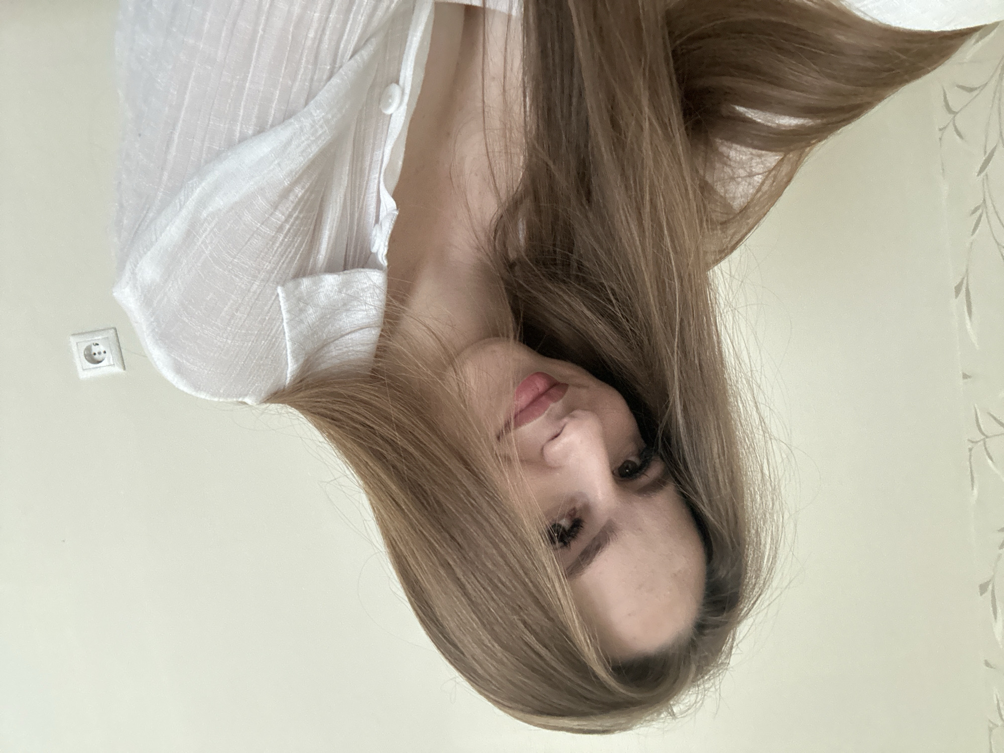 A young woman with long, light brown hair is captured in a close-up selfie, looking towards the camera with a slight smile. She is wearing a white collared shirt with buttons. The background is a plain, light-colored wall with a faint floral pattern on the left and an electrical outlet on the right. The lighting is soft, casting a gentle illumination on her face.