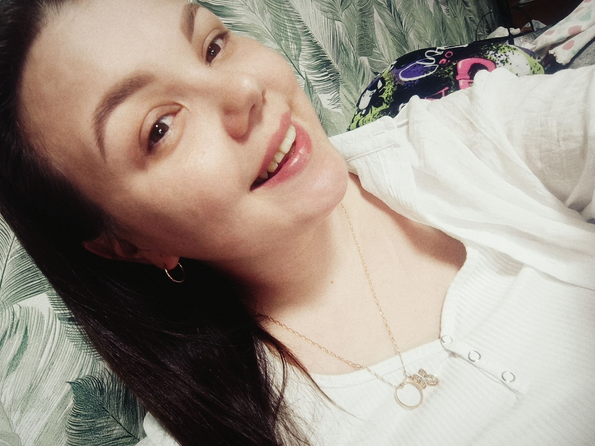 A close-up, slightly angled, indoor shot shows a light-skinned woman with dark hair smiling towards the camera. She has brown eyes and is wearing a small gold hoop earring in her left ear. She is also wearing a white ribbed top with small buttons and a gold chain necklace with a pendant featuring a butterfly and a circle. The background includes a green and white leaf-patterned wallpaper and some fabric with colorful abstract designs. The location cannot be determined from this photo.