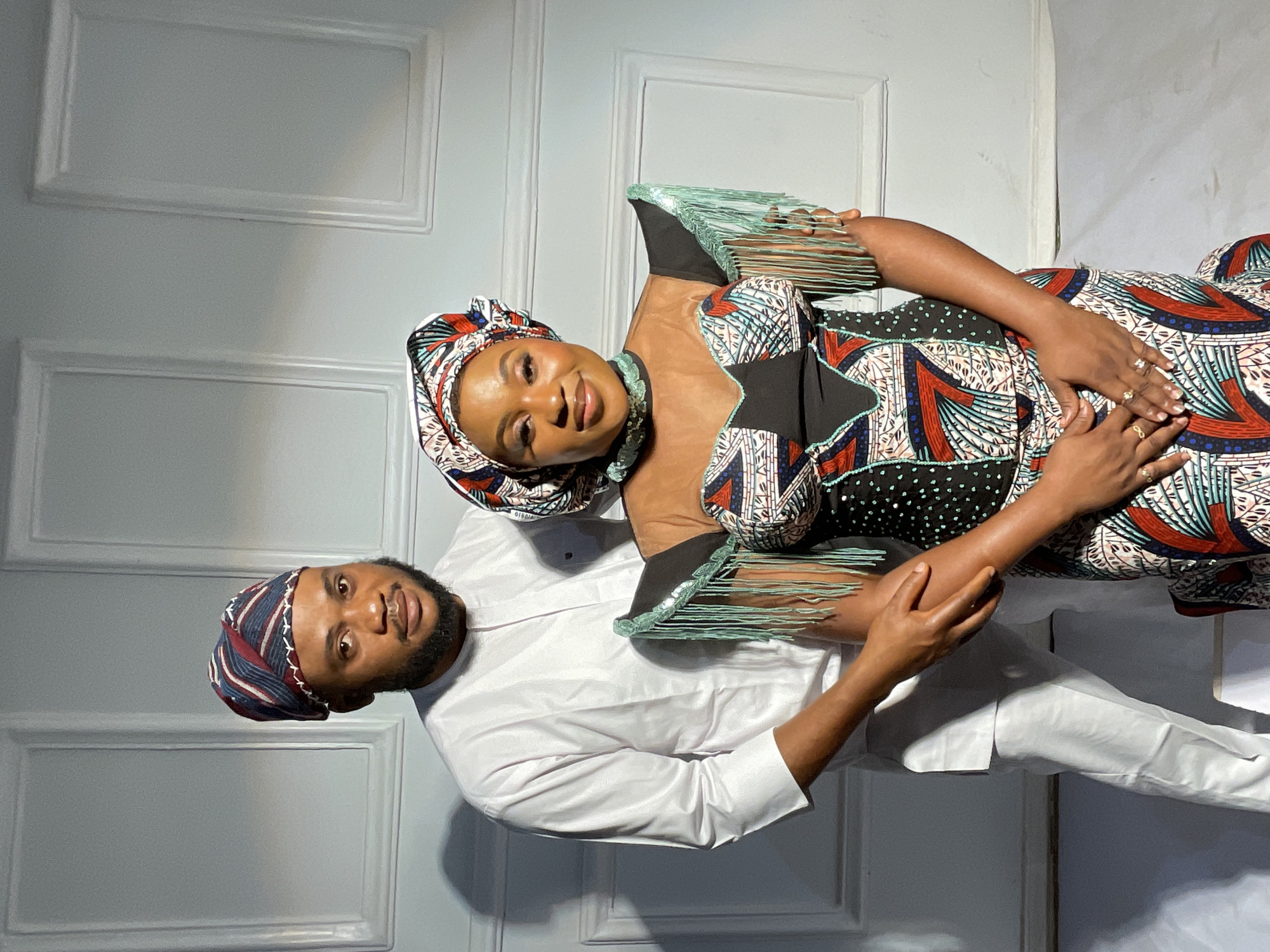 Two people, a man and a woman, pose for a portrait. The woman wears a patterned dress with fringe detailing and a matching headwrap. Her hands, adorned with rings, rest on her stomach. The man is in a white, long-sleeved shirt and pants, also wearing a matching headwrap. The background features a white paneled wall. The location is Barakin Akawo, Nigeria.