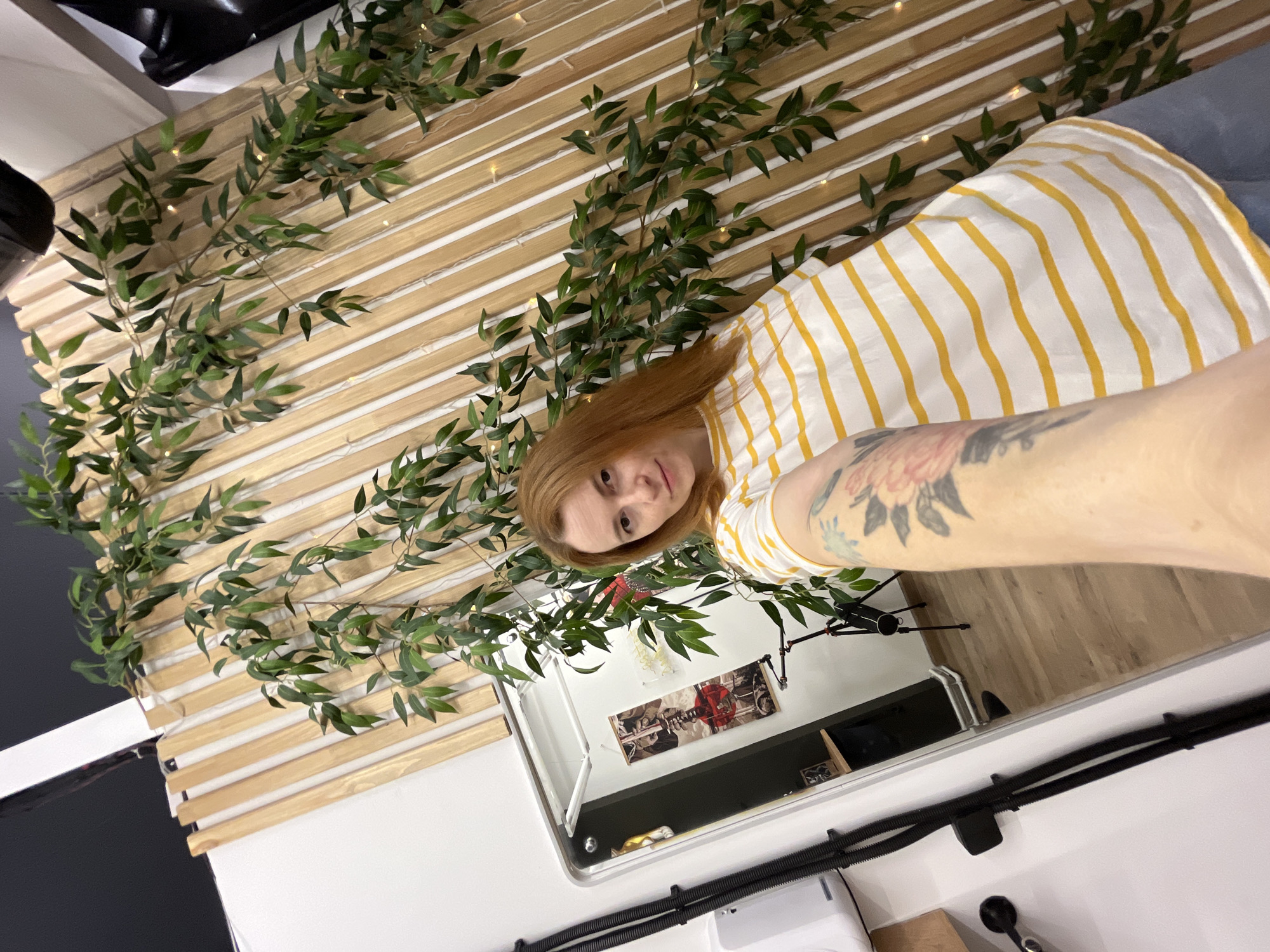 A woman with auburn hair is seen in a low-angle selfie. She is wearing a yellow and white striped shirt. Her left arm is extended towards the camera and features a tattoo on the inner forearm. Behind the woman, a light wooden wall with horizontal slats is decorated with green leafy vines. A framed mirror hangs below the wooden wall. The mirror reflects a desk with a microphone and various decorative items. Based on the location data, the image was taken in Chaikovsky, Russia.