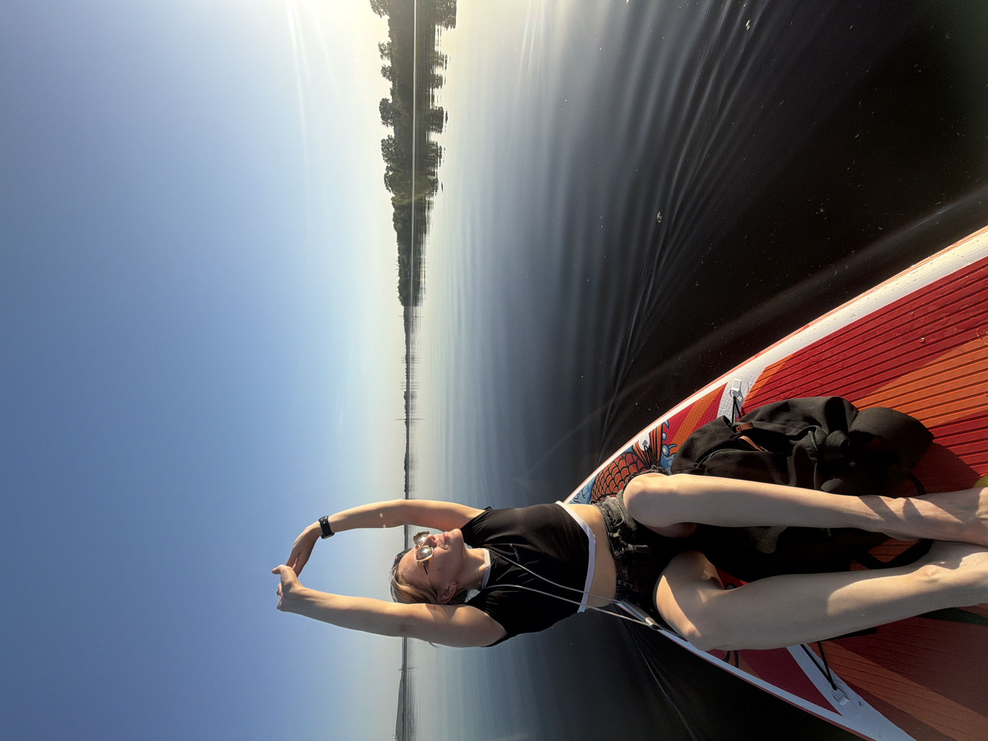 A woman is lying on a paddleboard on a body of water, seemingly a river or lake, possibly in the city of Noviy, Russia, based on the query. She is lying on her back with her arms raised above her head, stretching or relaxing. She's wearing a black cropped top and dark shorts, with sunglasses perched on her face. There is a black backpack near her feet on the paddleboard, which is red and orange in color. The water is calm with subtle ripples, reflecting the trees lining the opposite shore. The sky is blue and clear, with the sun shining brightly in the upper portion of the image.
