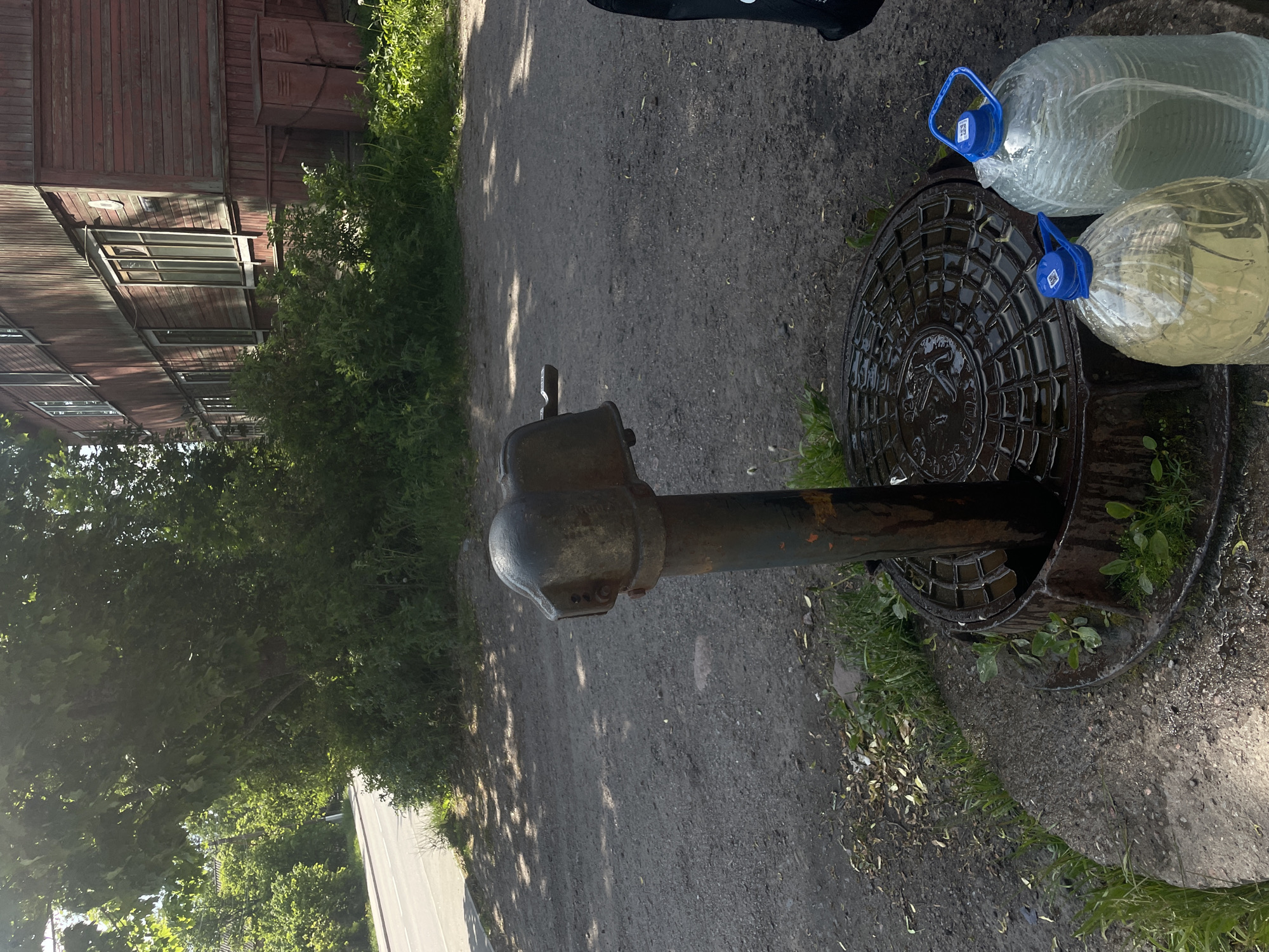 The image captures an outdoor scene in Priozersk, Russia, under clear daylight conditions. In the immediate foreground, a dark, weathered cast-iron manual water pump with a visible spout and operating handle stands centrally. The pump's base rests on a circular concrete slab, which in turn sits on a wet, circular metal manhole cover. The manhole cover exhibits a raised grid pattern and is covered in water, reflecting ambient light, indicating recent activity at the pump. Adjacent to the pump, positioned on the wet manhole cover, are two large, clear plastic bottles, each with a blue plastic handle and cap. One bottle contains a clear liquid, while the other holds a pale yellow liquid. Both bottles show external condensation or water droplets.
The ground around the pump is composed of dark greyish-brown dirt and loose gravel, with sparse patches of green grass and weeds growing through it. In the midground to the left, a paved asphalt road with a single white lane marking is visible. The midground and background feature dense green vegetation, including bushes and trees. To the right in the background, a two-story wooden building with faded reddish-brown plank siding is prominent. The building has multiple windows with light-colored frames. Below the building's facade, several rusty, reddish-brown metal utility boxes or cabinets are attached to the lower wall. The scene is illuminated by direct sunlight, casting distinct shadows across the ground and highlighting the textures of the various objects. No human presence is observed.