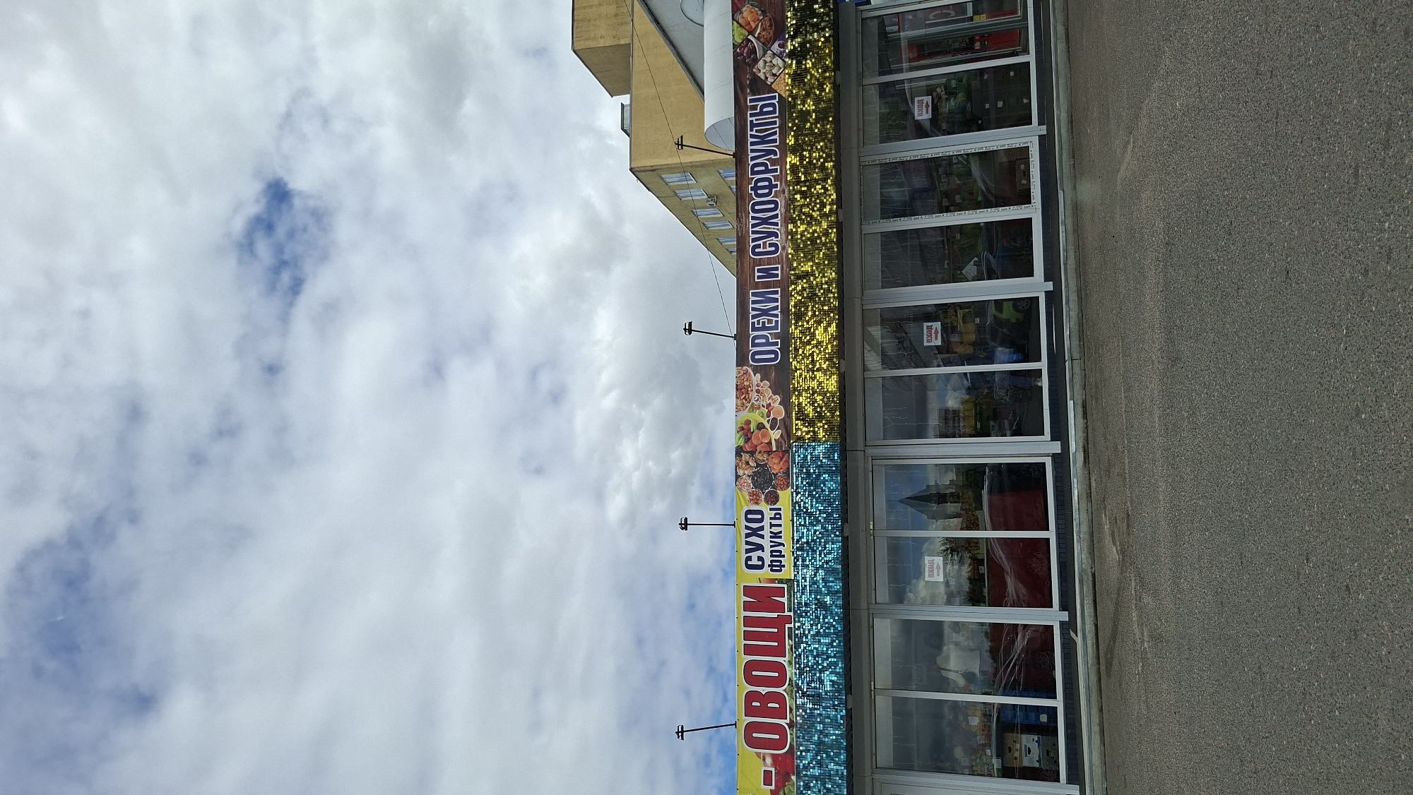 The image shows the exterior of a store in Maladzyechna, Belarus, under a cloudy sky. A large banner across the top of the building advertises "ОВОЩИ" (vegetables) and "ОРЕХИ И СУХОФРУКТЫ" (nuts and dried fruits). The banner is colorful, featuring images of produce and nuts, and is decorated with blue and yellow sequined sections. Below the banner, the store has large glass windows, through which some interior shelves and products are visible, along with reflections of the surroundings. Red "ВХОД" (entrance) signs are placed above some of the windows. A paved area, likely a parking lot or sidewalk, is in the foreground. The building itself is modern with a yellow facade visible on the right side.