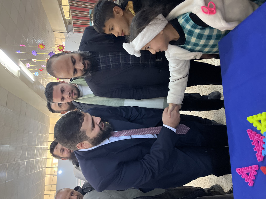 The image depicts an indoor scene in Kafr Batna, Syria, featuring seven individuals. A bearded adult male, wearing a dark suit, white shirt, and patterned tie, is shaking hands with a young girl. The girl, dressed in a light-colored top, plaid dress, and a white headband with a bow, is smiling and looking at the man. To the girl's right, a young boy in a dark jacket observes the handshake. Behind the girl, a bearded adult male in a dark jacket and plaid shirt also observes the interaction. Three other adult males are visible in the background: one partially visible to the far left, another bearded individual centrally behind the handshake, and a third male to the right of the central figure wearing a light green scarf and a dark jacket. In the immediate foreground, a blue table displays several brightly colored plastic geometric shapes (pink, blue, yellow triangles). The environment consists of white walls, fluorescent ceiling lights, and colorful paper flower decorations suspended from the ceiling. A striped fabric is visible on the far right background.