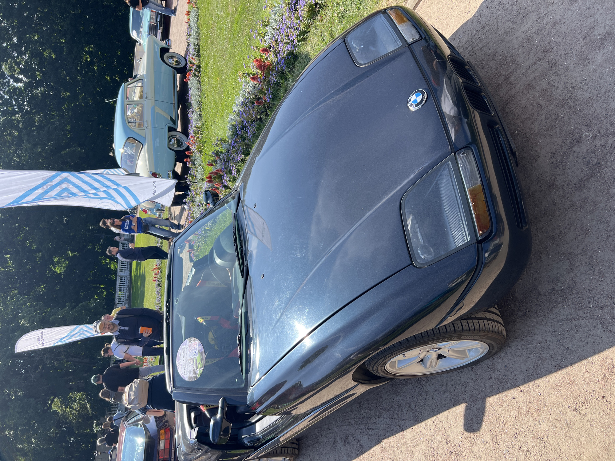 A dark blue convertible BMW Z1 is parked on a gravel path, facing right. The car has a black interior and racing harnesses visible through the open doors. The BMW emblem is on the hood. Behind the car, a grassy area is filled with flower beds. People are milling around, and several large flags with abstract blue and white designs are planted in the ground. A light blue and white vintage car is parked in the background, along with other vehicles and trees. The scene appears to be an outdoor car show or event, possibly in a park.