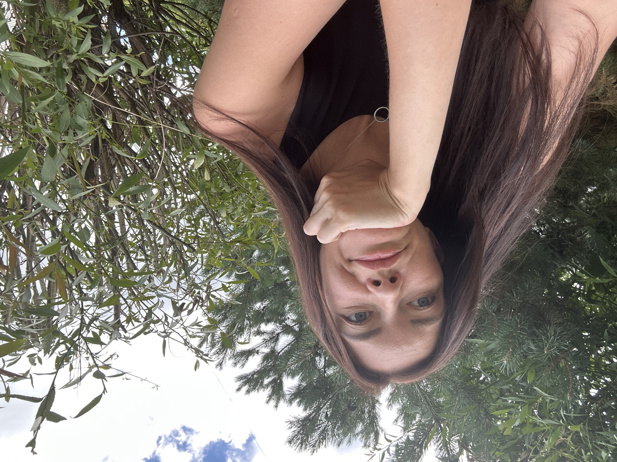 A woman with long, dark brown hair is taking a selfie outdoors, leaning her chin on her hand. She is wearing a black sleeveless top and a silver pendant necklace. Behind her, there are lush green trees and a cloudy sky. The image appears to be taken in a natural setting, possibly a garden or park, with no visible urban structures or elements of the city of Даниловка (алтайский), Russia. The lighting suggests it is daytime.