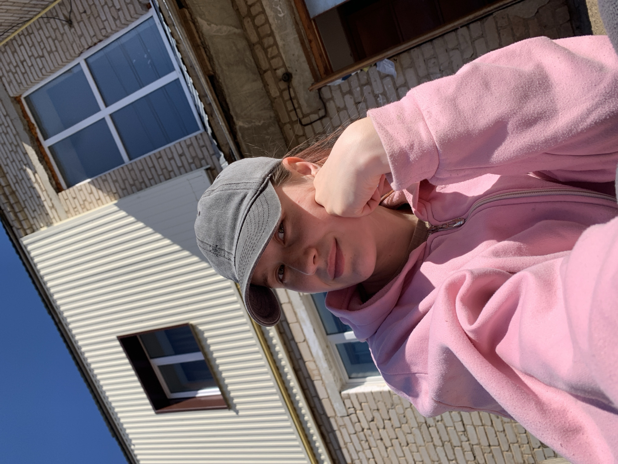 A young person with fair skin and light brown hair is wearing a gray baseball cap and a pink hooded sweatshirt. The individual is holding their fist near their cheek. The person is situated outside, in front of two buildings. One building is covered in light-colored siding, and another is built with white brick. A clear blue sky is visible in the upper left corner. The location is Фоки, Russia.