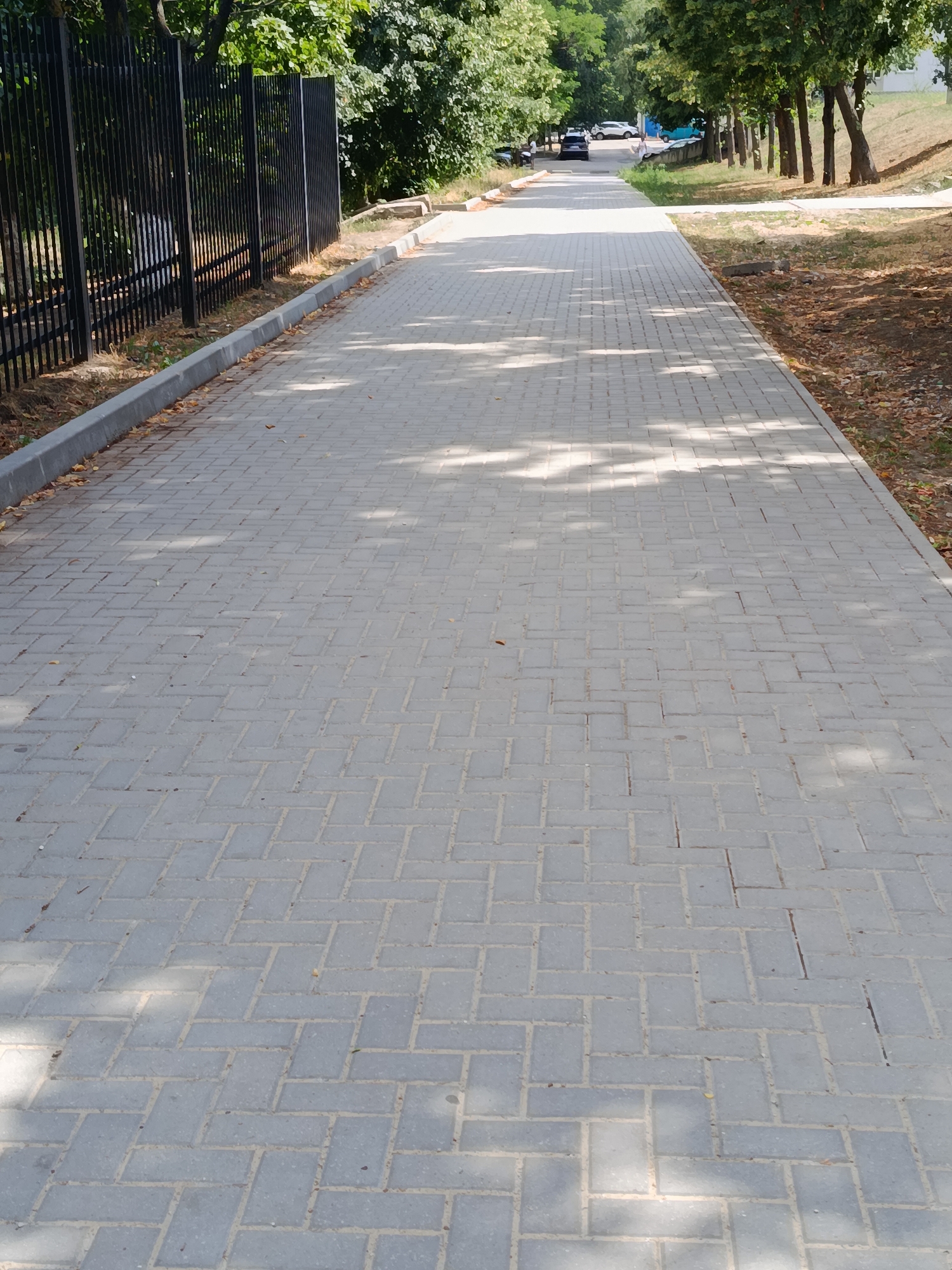 The image shows a paved walkway lined with a black metal fence on the left and trees on the right. The walkway is made of gray rectangular pavers laid in a herringbone pattern. Sunlight filters through the leaves of the trees, casting dappled shadows on the path. In the distance, several cars are parked along the road, and a few people can be seen walking. The scene appears to be in a park or a residential area in Chișinău, Moldova. The overall impression is one of a quiet, sunny day in an urban green space.