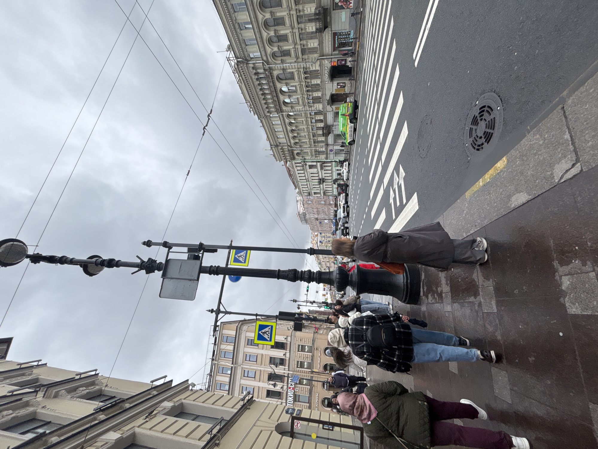 The image shows a street scene in Saint Petersburg, Russia. People are standing on a wet sidewalk next to a street with a pedestrian crosswalk. Several buildings line the street. A light fixture and wires span across the street. A crosswalk sign is visible. The sky is overcast.