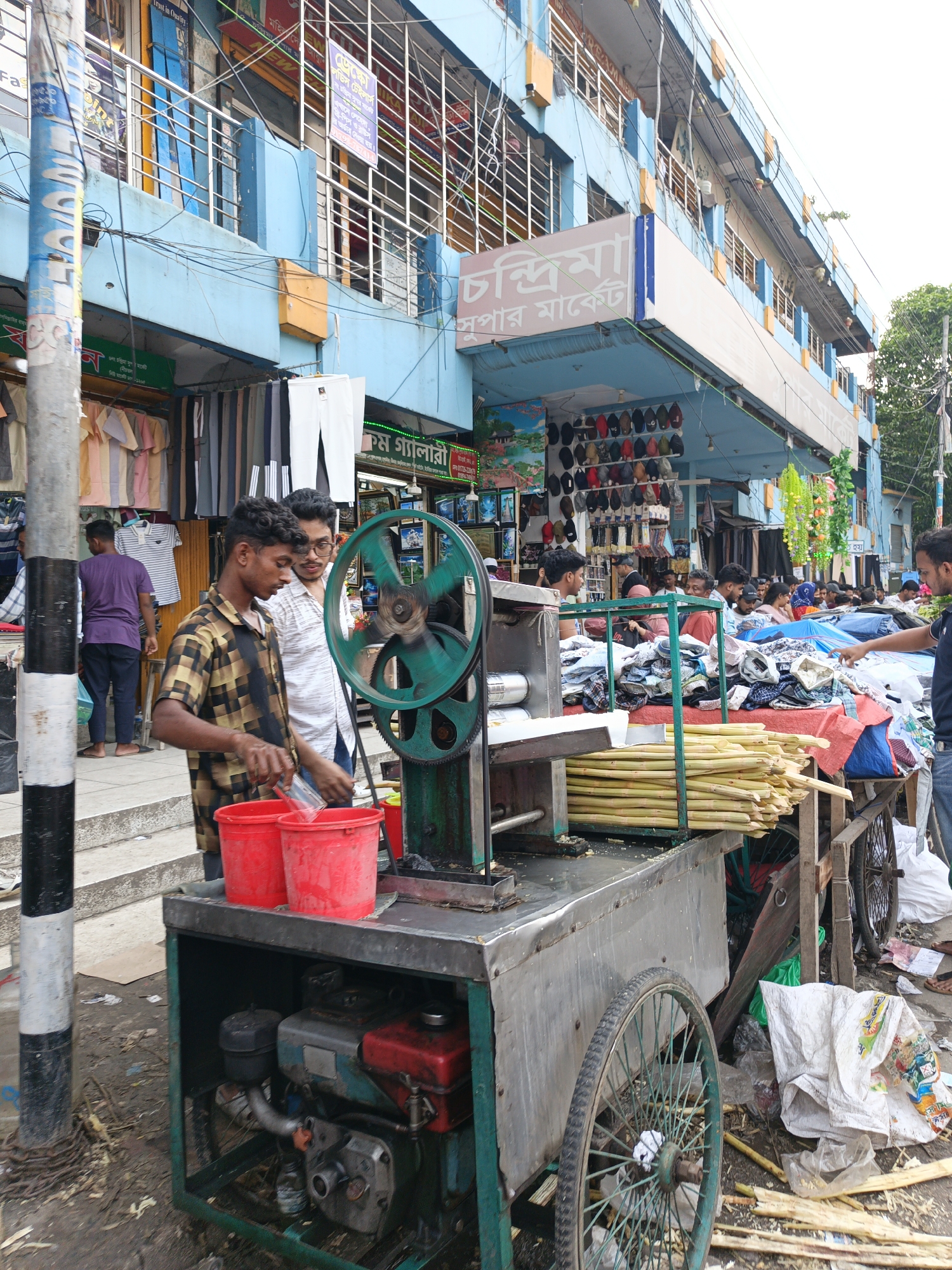 Here is a detailed description of the image:
**Scene Overview:**
The image depicts a bustling street scene, likely in a commercial area of an urban setting. A primary focus is a mobile sugarcane juice vending cart, operated by two men. The backdrop reveals multi-story buildings with shops and signs, indicative of a market or commercial district.
**People:**
* **Vendor 1:** A young man with dark curly hair stands at the cart, wearing a gold and black checkered shirt. He is pouring liquid from a glass into a red bucket.
* **Vendor 2:** Another young man, wearing glasses and a white shirt, stands beside the first vendor.
* **Customers/Pedestrians:** A number of people are visible in the background, browsing goods at stalls, walking, and generally going about their business. A few are near the juice cart, possibly customers.
**Objects:**
* **Sugarcane Juice Cart:** A homemade cart with a motorized sugarcane juice extracting machine, painted in green. The machine has a prominent green flywheel. Two red buckets are positioned on the cart to collect the juice. Several stalks of sugarcane are bundled and stored on the cart. The cart has bicycle wheels, suggesting it's mobile.
* **Sugarcane:** Bundles of sugarcane stalks, the primary ingredient for the juice being sold, are visible on the cart.
* **Merchandise/Stalls:** In the background, there are clothes displayed for sale, indicating a clothing market.
* **Buildings:** The buildings in the background appear to be commercial establishments, possibly a market complex. They have multiple levels, balconies, and various signs in a non-English script.
* **Street Elements:** A black and white striped pole, possibly a utility pole, is in the foreground. There is debris and litter on the street.
**Actions and Interactions:**
* The vendors are actively operating the sugarcane juice cart. One is pouring juice, likely serving a customer.
* People are browsing merchandise at the stalls in the background, suggesting active commercial activity.
**Environment:**
* **Location:** This is most likely an urban street in an area with active commerce. Buildings and multiple people on the street support this assessment.
* **Light:** The lighting is bright, suggesting it is daytime, but not harsh, potentially indicating partial cloud cover.
**Additional details**
The text on the signs appears to be in Bengali.
The cart appears to have a small engine to power the sugarcane juicer.
The environment is somewhat cluttered with trash and other debris.
---
