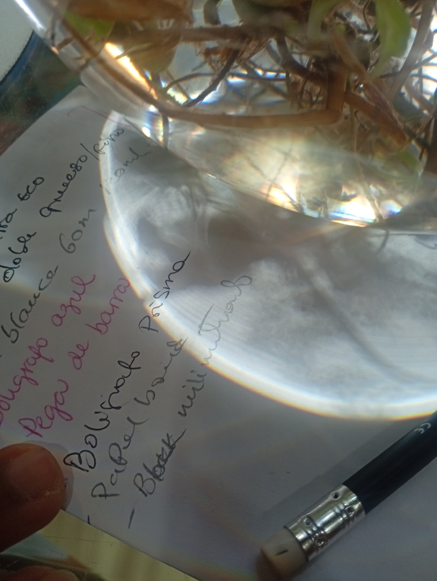 The image shows a close-up of a handwritten list on a piece of white paper, partially obscured by a finger. The paper is placed on a surface that appears to be a table, and it is illuminated by a bright light source, possibly sunlight, which casts reflections and refractions on the paper and a glass container in the background. The handwriting is in black, pink, and red ink.
The visible text includes:
- "eco"
- "doble queso/fino"
- "blauce Gorn"
- "cougtafo azul"
- "Pega de barra"
- "- Boligrafo Prisma"
- "- Papel bond"
- "- Block mill"
In the background, there is a glass container with plant roots and possibly water. A pencil with an eraser is also visible at the bottom right corner of the image. The overall scene suggests a domestic or creative environment, possibly a desk or workspace. There is no indication of the city of Juan Griego, Venezuela, within the image itself.