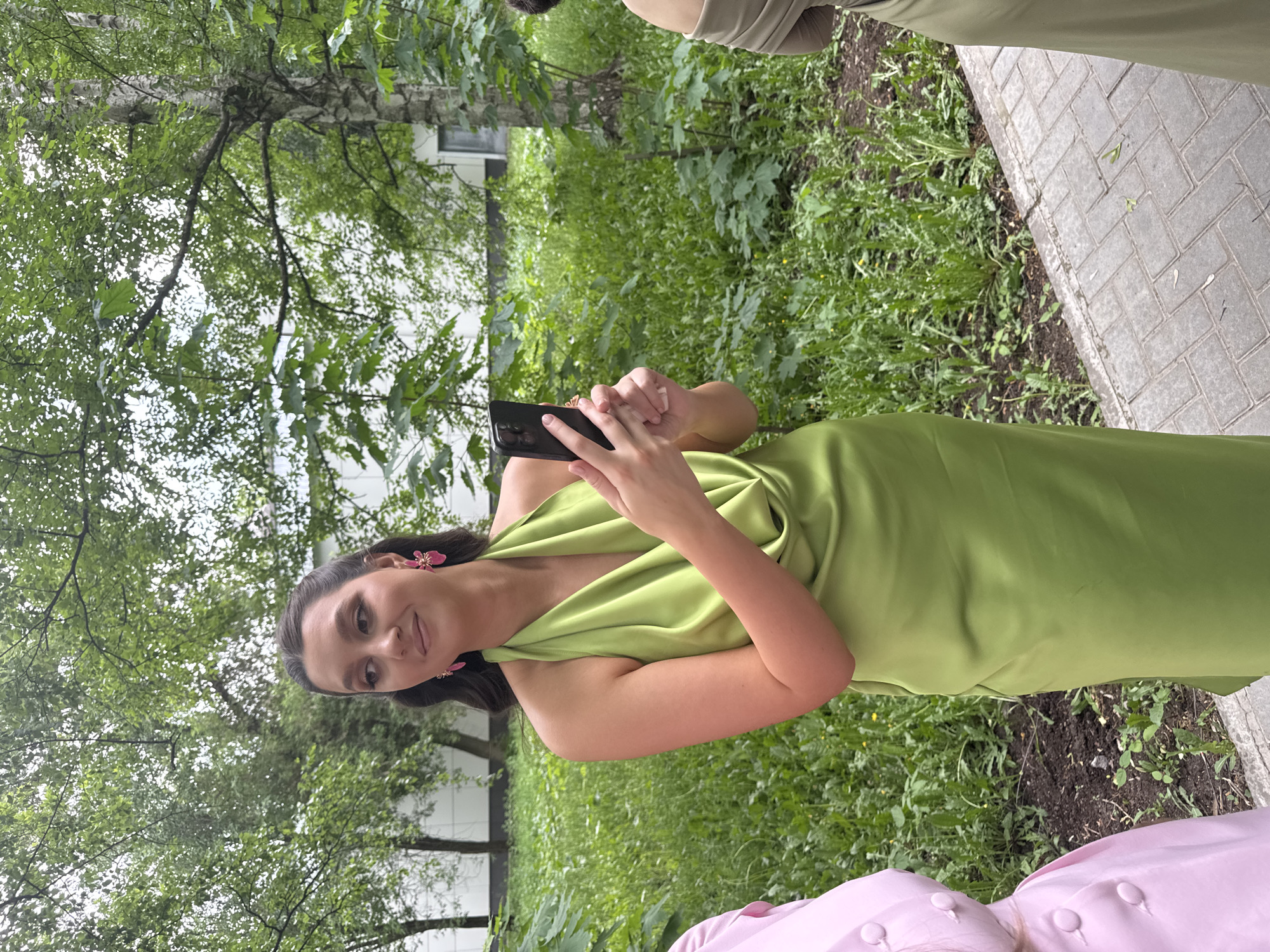 A young woman in a long, green dress stands outside, holding a smartphone in both hands. She is facing the camera, with a slight smile on her face. She is wearing pink flower-shaped earrings. The background consists of green foliage and trees, suggesting a park or garden setting. A sidewalk is visible on the right edge of the frame, with greenery growing between the sidewalk and the street. The location is Perm, Russia. Another person can be seen on both the top and bottom of the frame, each wearing a gown in a different shade of green and pink, respectively.
