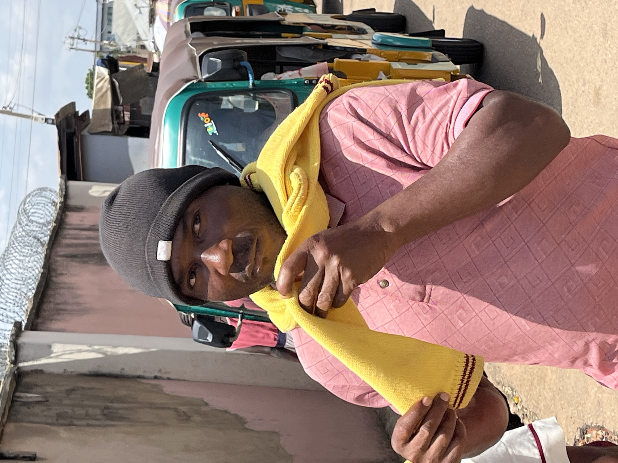 An outdoor, eye-level medium shot captures a man in a black beanie and a pink patterned shirt standing in what appears to be a street or thoroughfare in Jos, Nigeria. He is holding a yellow scarf with both hands, wrapping it around his neck and chest. Behind him, a line of three green and yellow auto-rickshaws are parked or moving slowly. The background includes a pink wall topped with barbed wire, buildings, and a cloudy sky. The man's gaze is directed forward, and his expression is neutral. The lighting suggests it is daytime.
