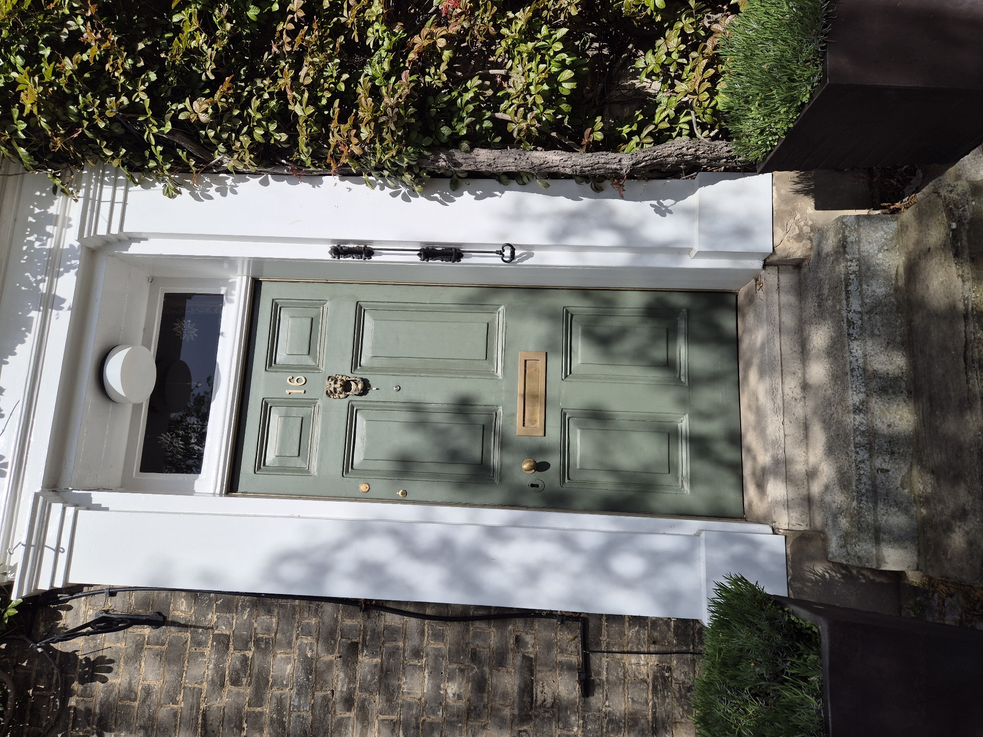The image shows a front door in West Suffolk, United Kingdom. The door is painted a muted green and has six rectangular panels, a brass letter slot, a lion's head door knocker, and the number "91" above it. A small window is on the left side of the door. The doorframe is white. Above the doorframe is a dark green hedge. To the right of the door is a stone step, and a dark planter with green plants is visible next to the doorframe. The ground in front of the door is paved with dark brick.
